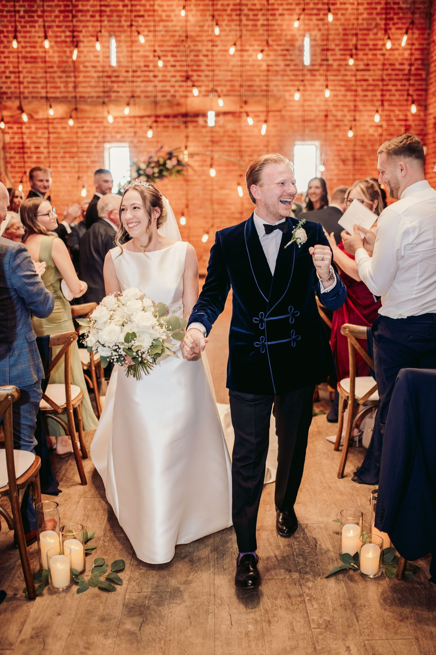 Wedding guests dance and celebrate at a rustic brick venue illuminated by warm string lights and candles.