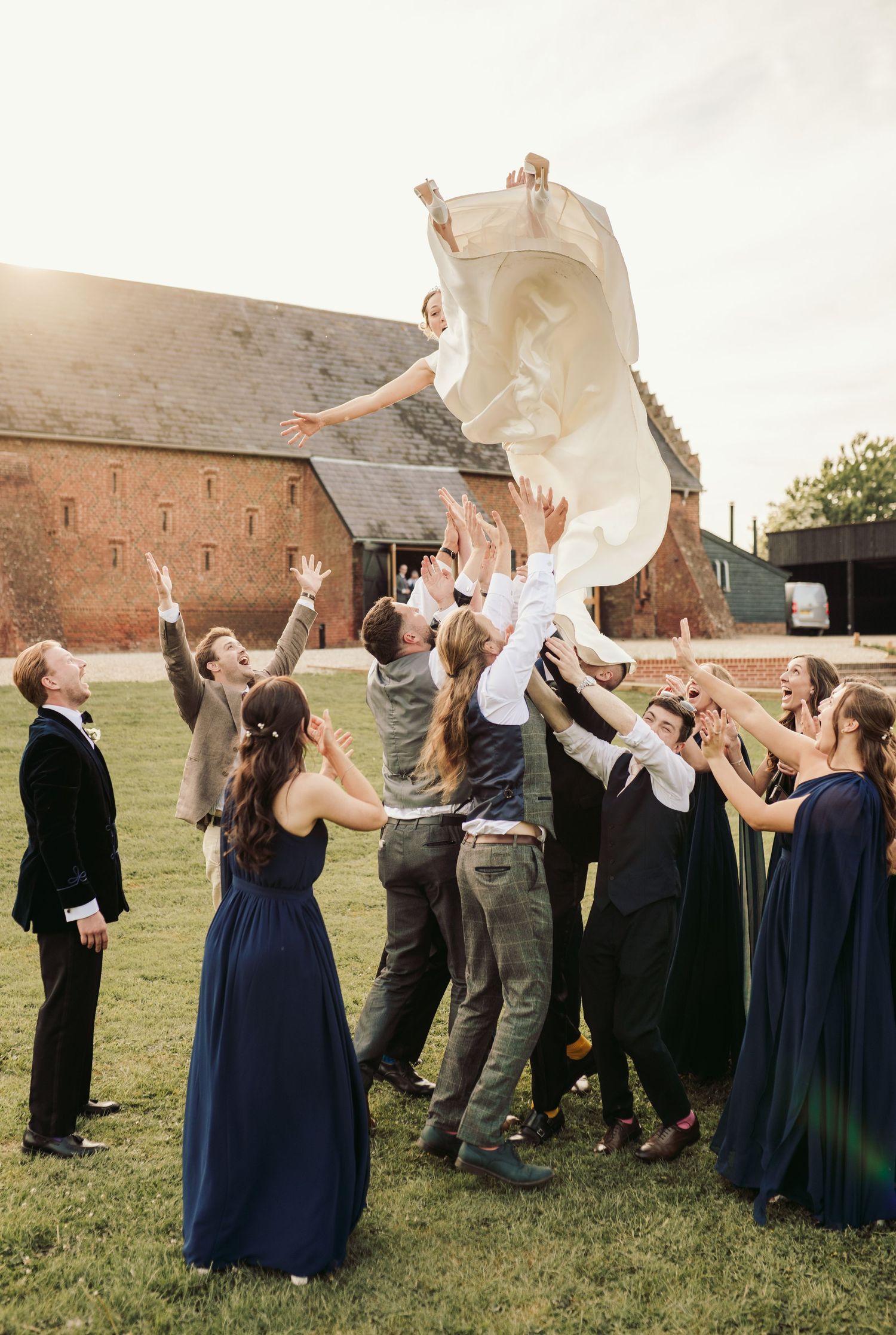 A group in formal attire performs a celebratory blanket toss on a grassy lawn near historic brick buildings at sunset.