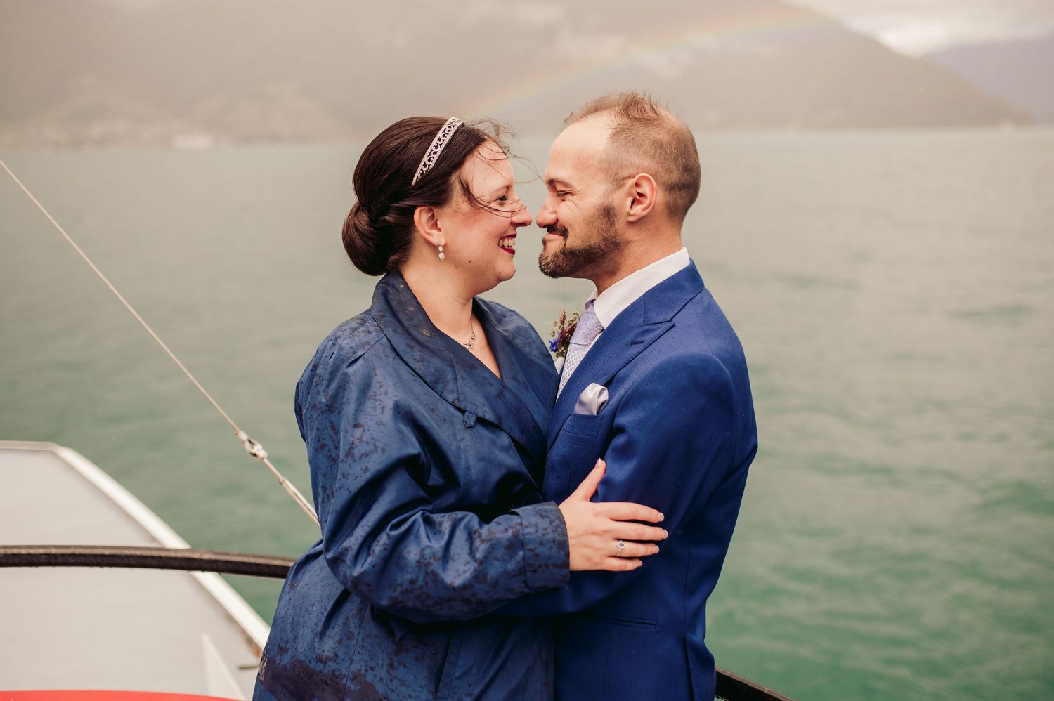 A couple in blue coats shares a romantic moment while standing on a boat with misty waters in the background.Spiez switzerland destination