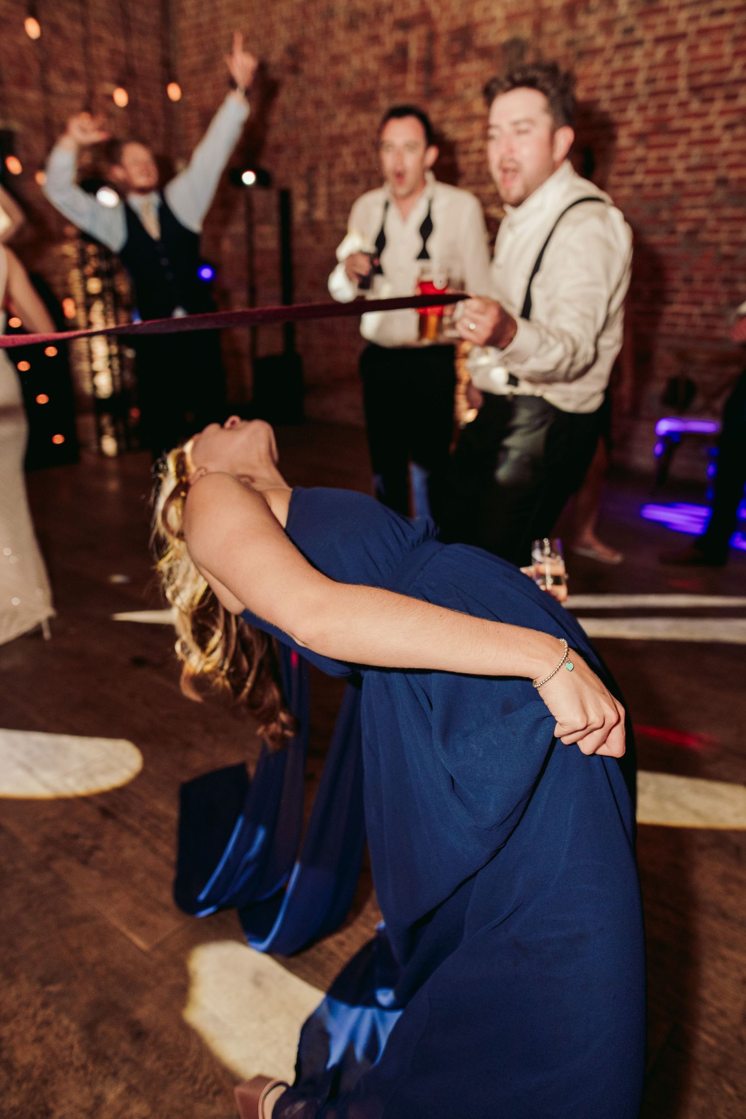 Wedding guests dance and celebrate on a rustic brick-walled dance floor illuminated by ambient lighting.