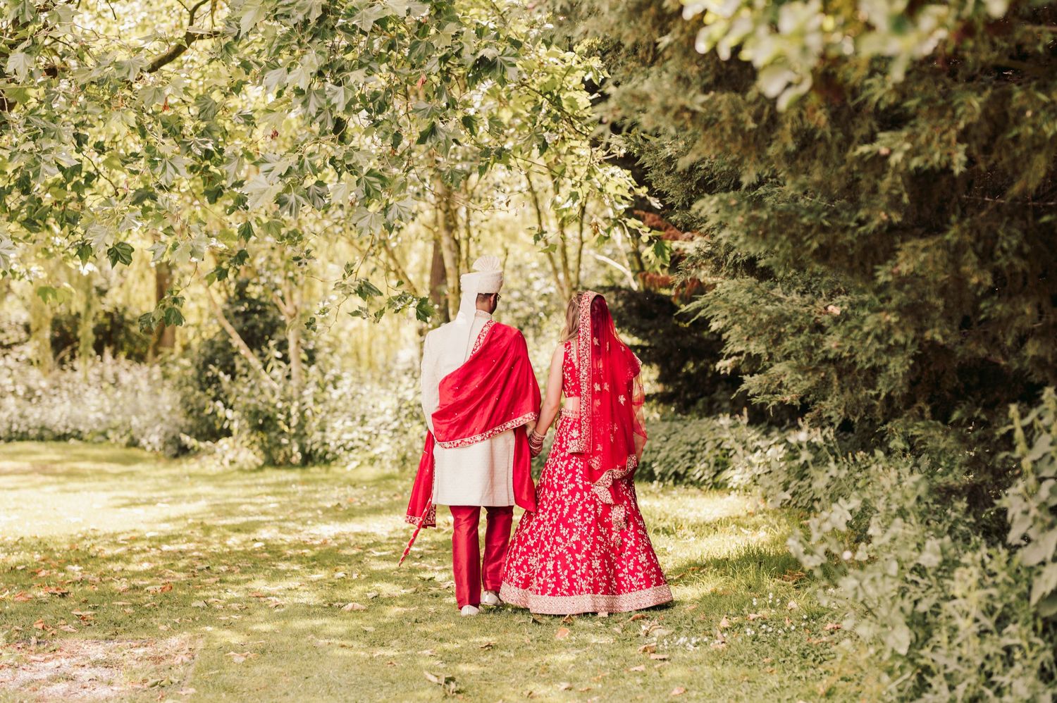 A couple in bright red traditional wedding attire walking together on a garden path surrounded by greenery.