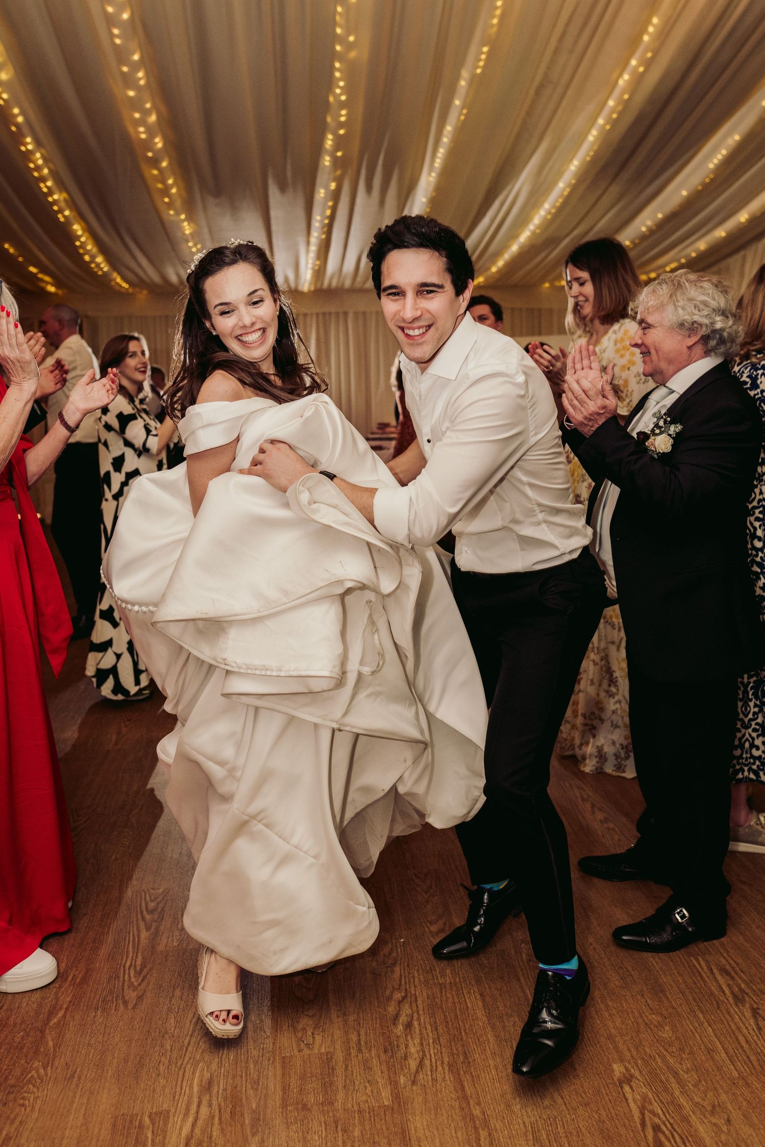 Couple performing energetic dance move during wedding reception with guests cheering in background.