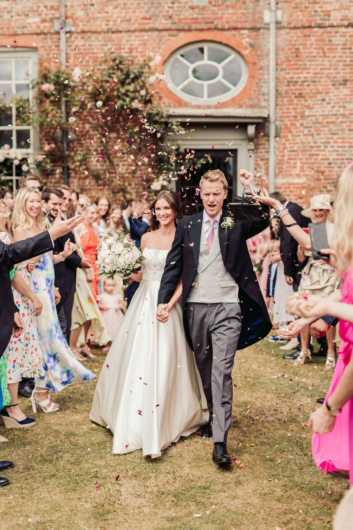 Wedding party and guests celebrate with confetti toss outside brick mansion.