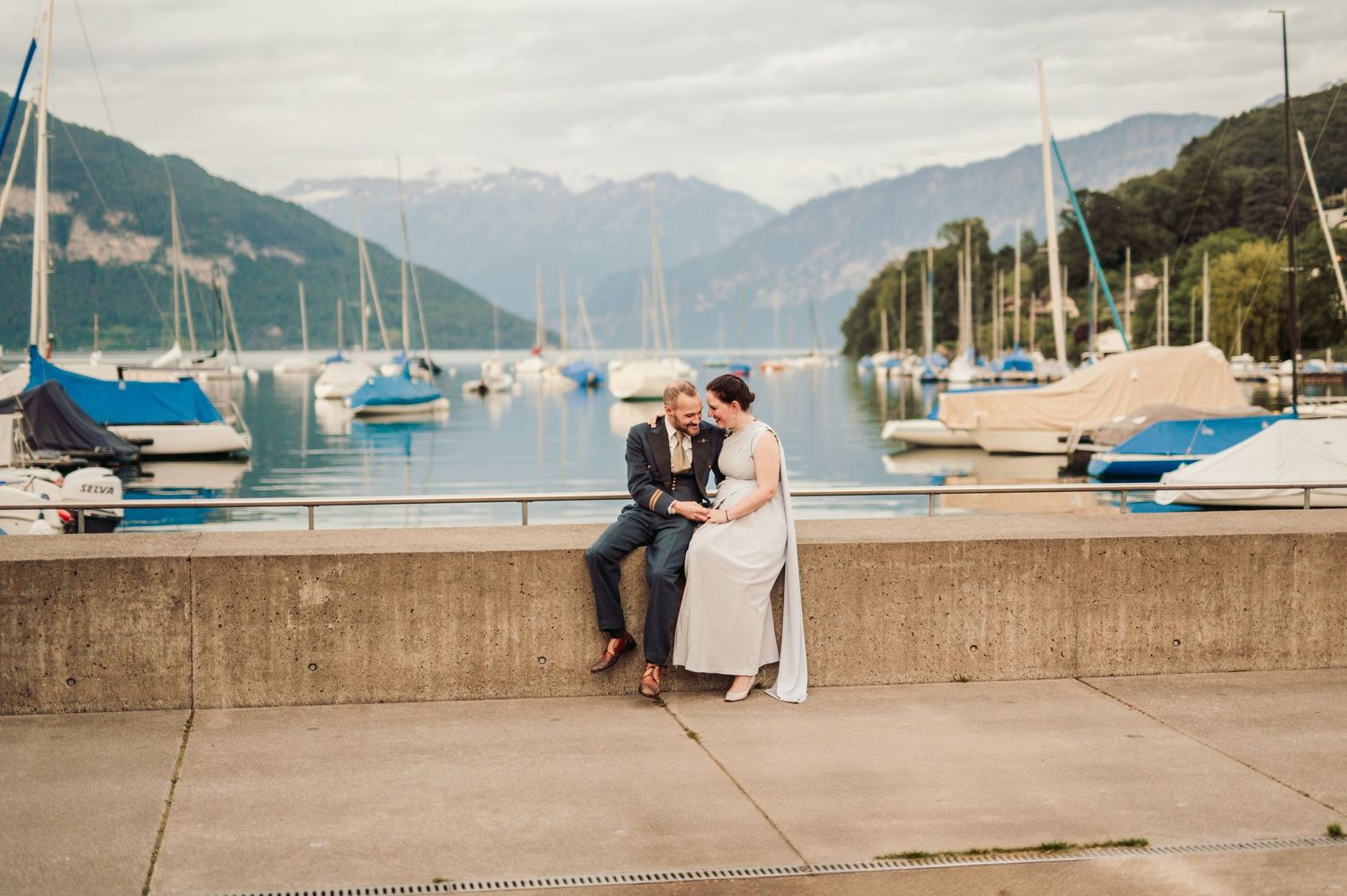Two people sit together on a marina wall overlooking a peaceful mountain lake filled with sailboats and yachts.Spiez switzerland destination