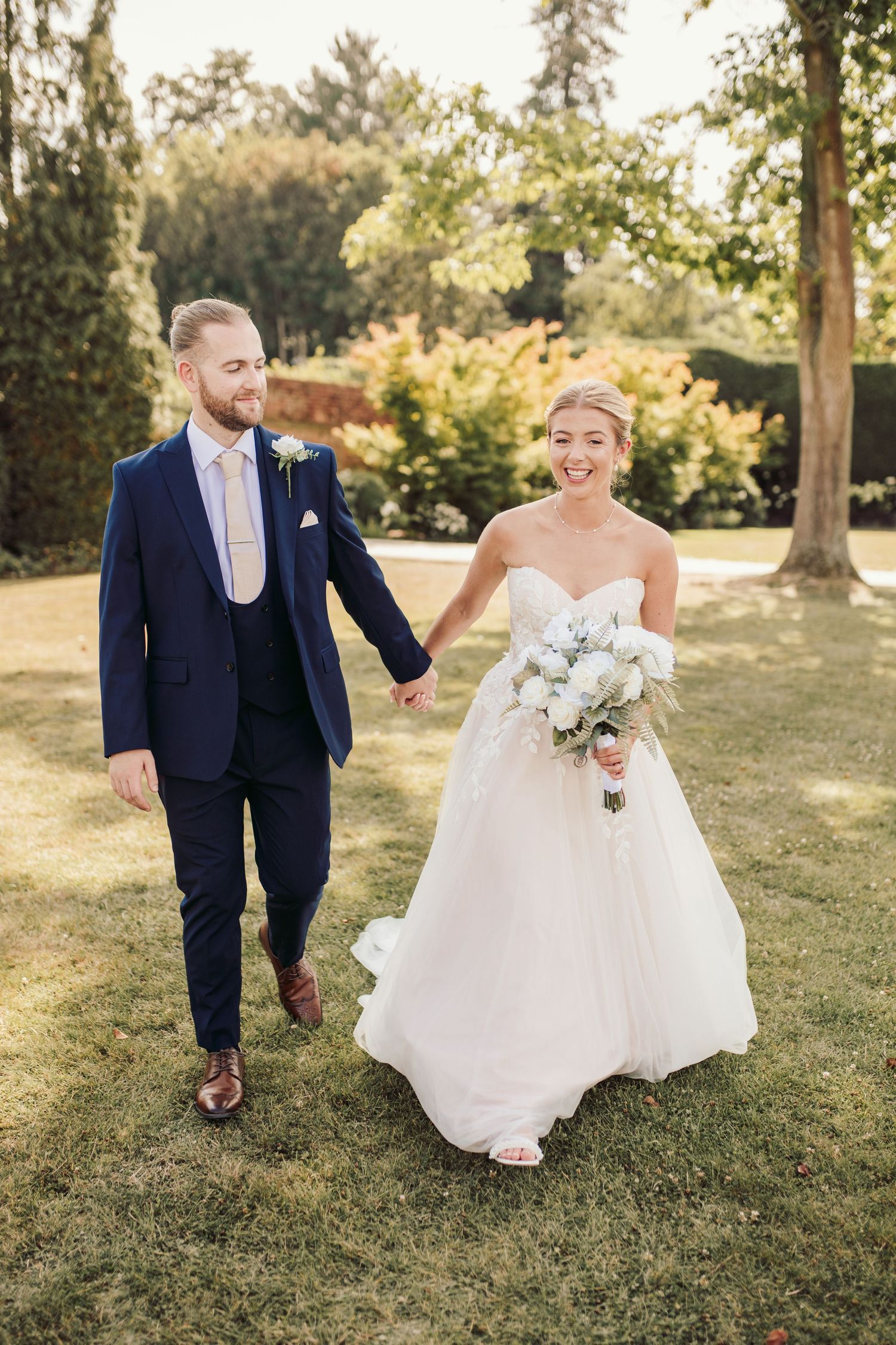 Couple running playfully across sunny lawn in formal wedding attire.