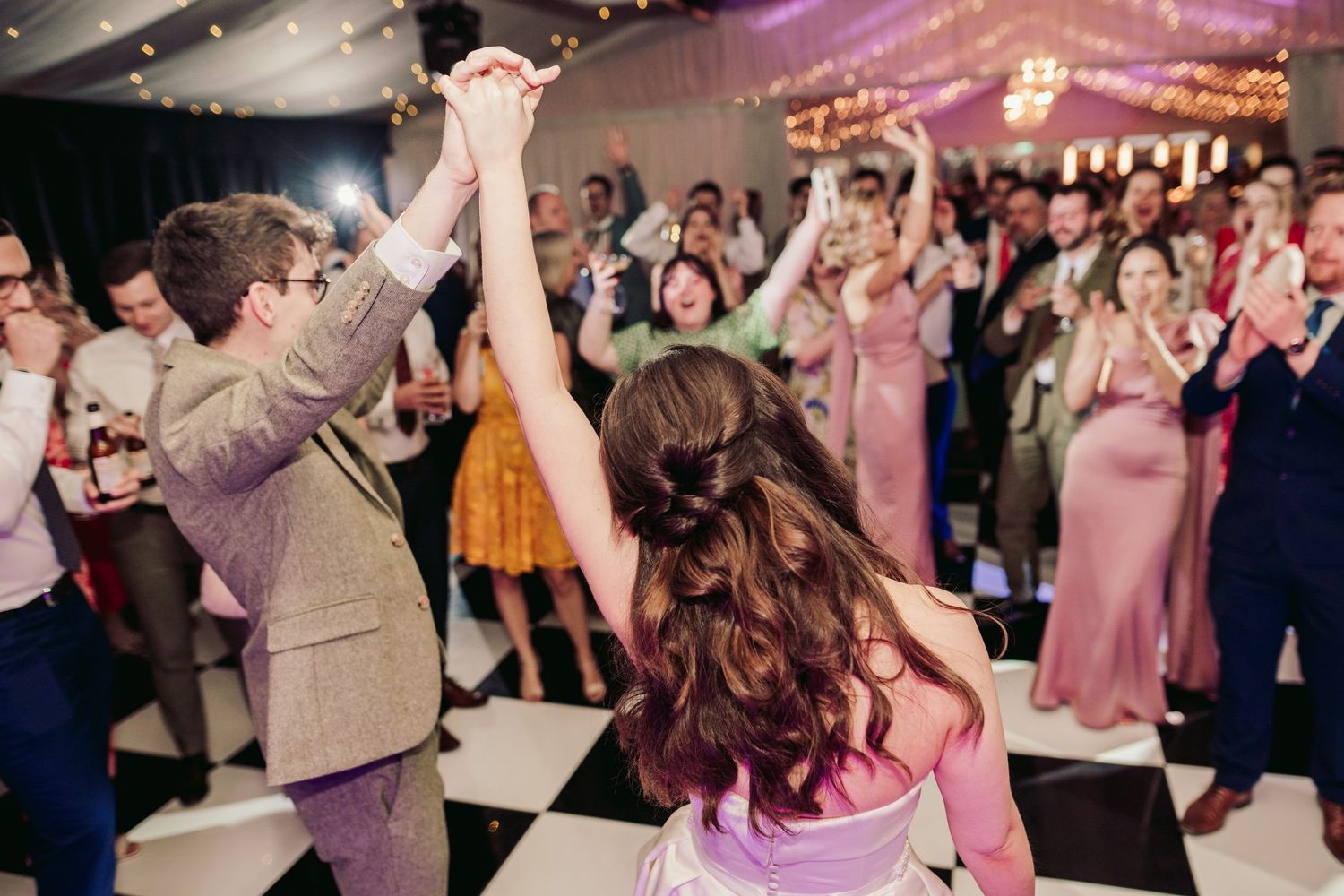 Wedding guests dancing with raised arms on checkered dance floor under twinkling lights.