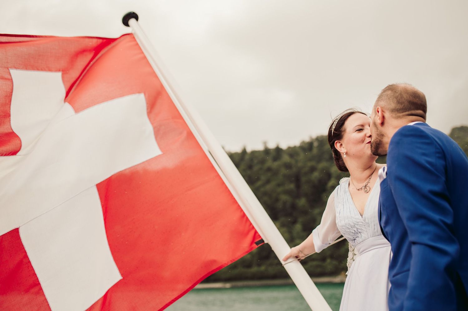 A couple shares a romantic moment by a lake with the Swiss flag waving in the foreground. Spiez switzerland destination wedding