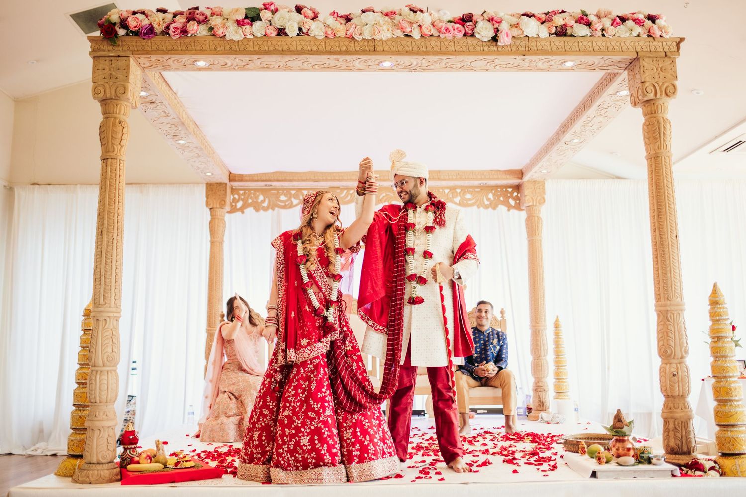 Traditional Indian wedding ceremony taking place under ornate gold mandap with red floral decorations and rose petals.
