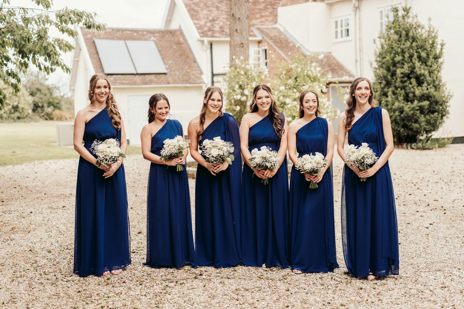 A row of bridesmaids in navy blue dresses stand together holding bouquets at an outdoor wedding venue.