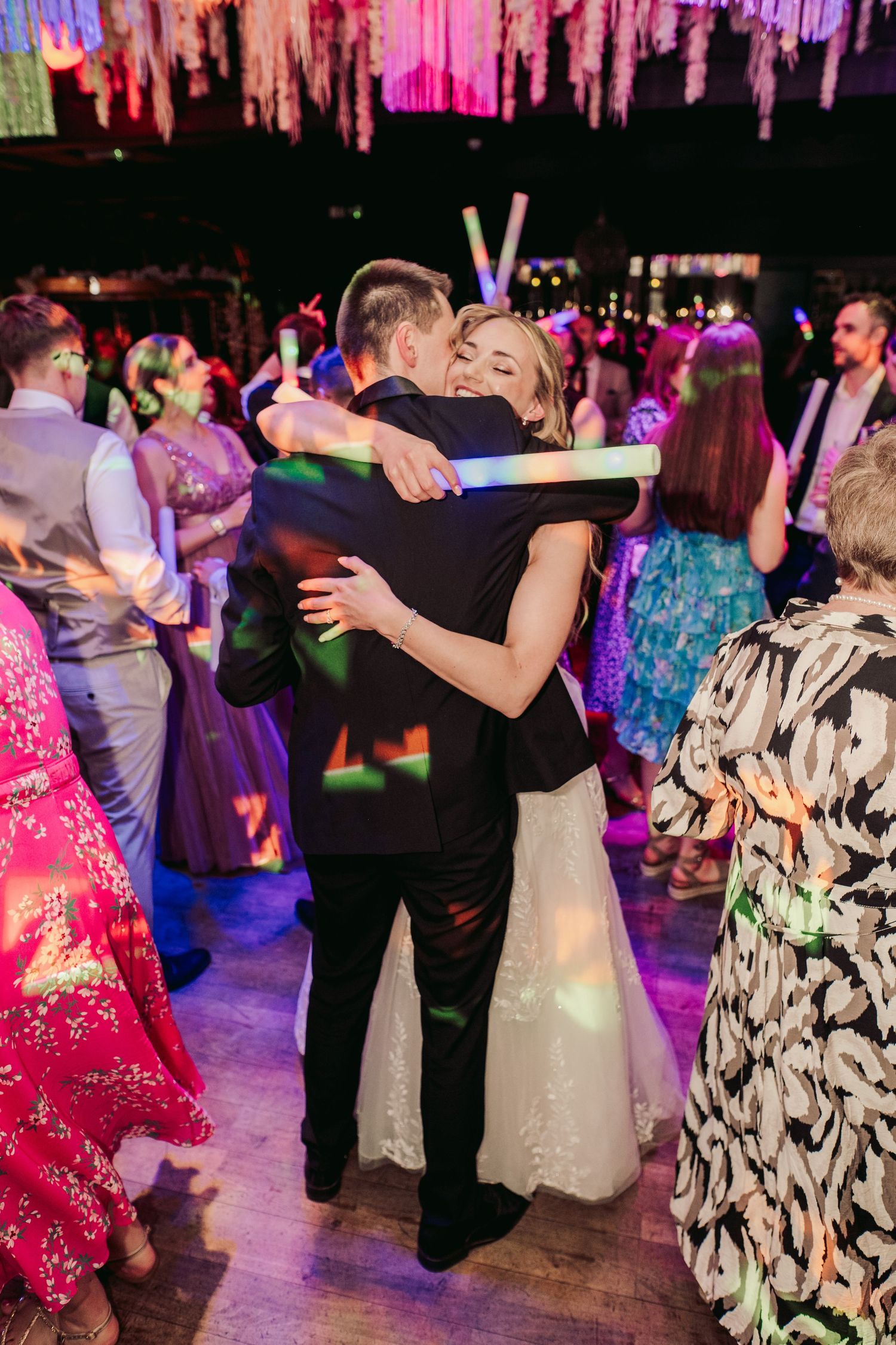 Couple embraces on a dance floor under pink lighting while guests dance at a wedding reception.