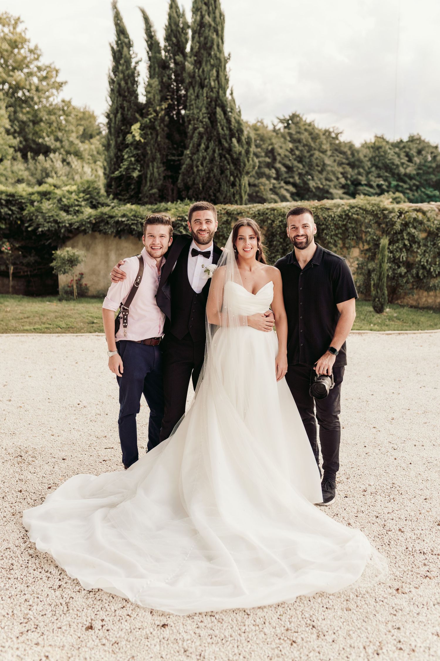 Wedding party posing outdoors with flowing white dress against cypress trees.
