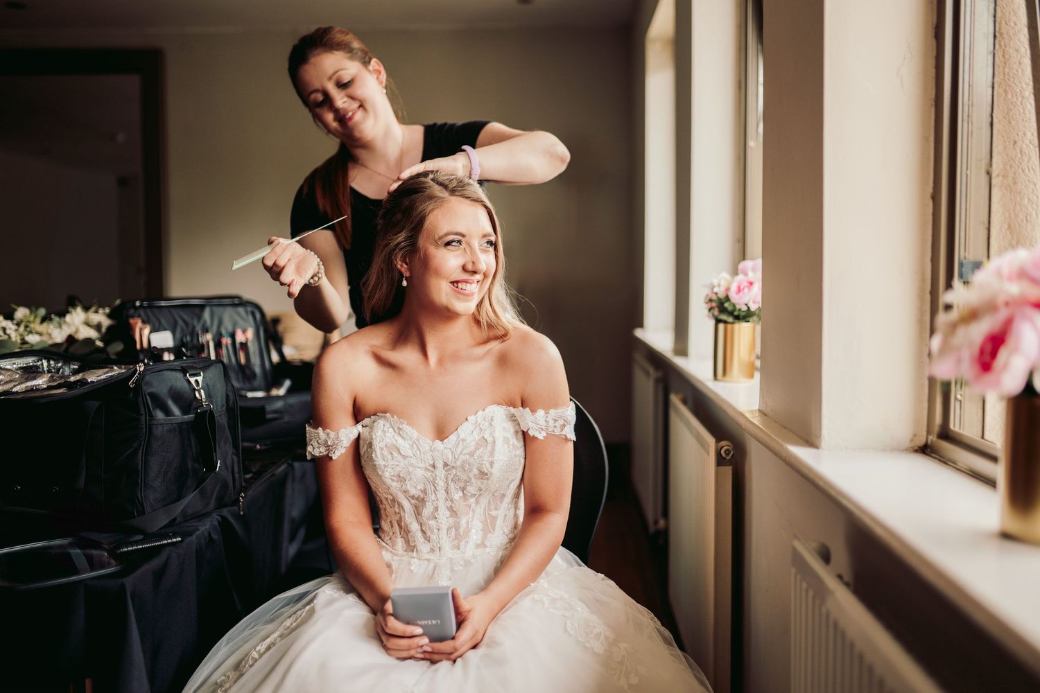 A hairstylist works on styling a bride's hair as she sits in her white wedding gown by a window.