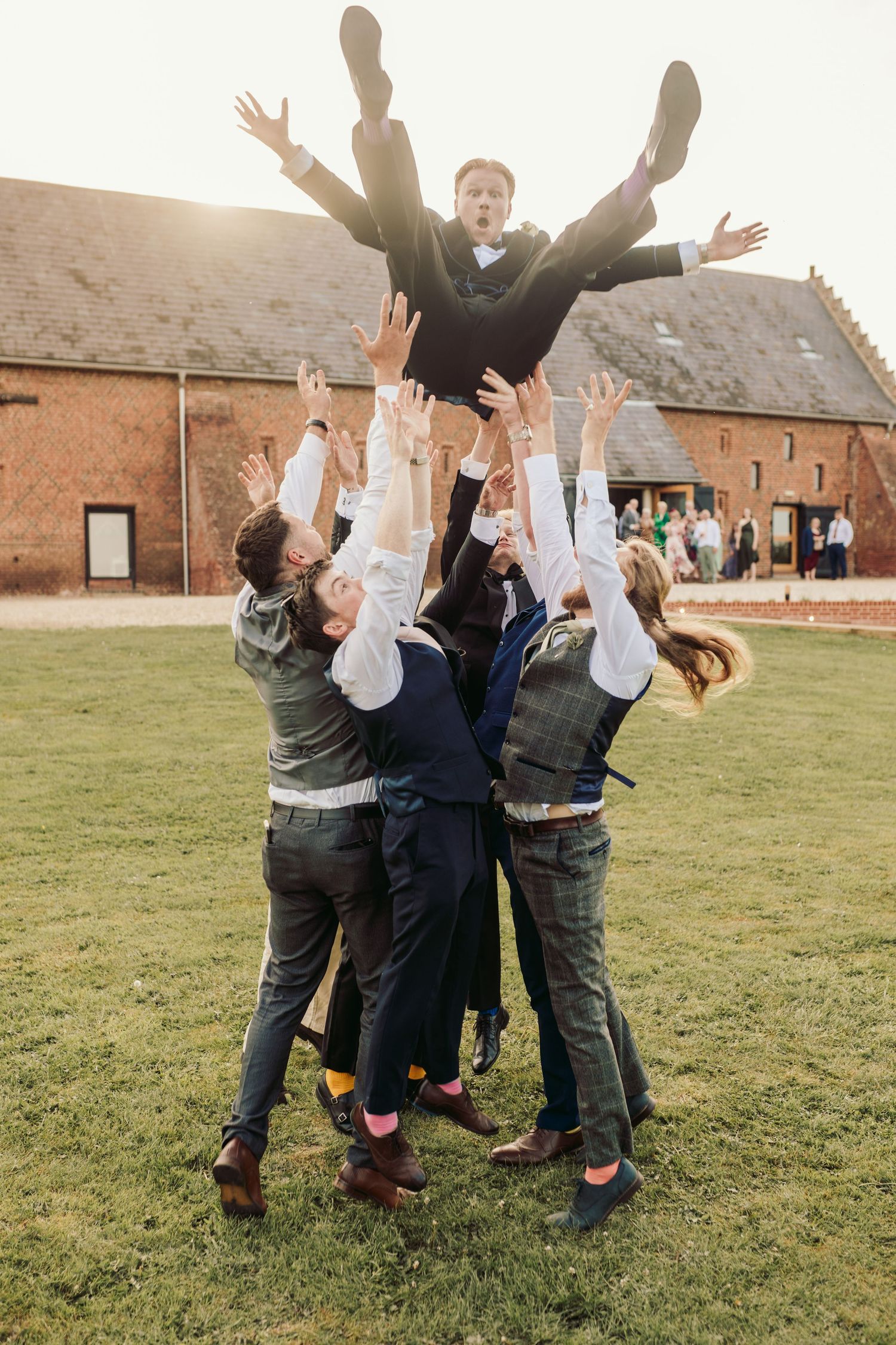 Group of people jumping and celebrating together on grassy field outside historic brick buildings.