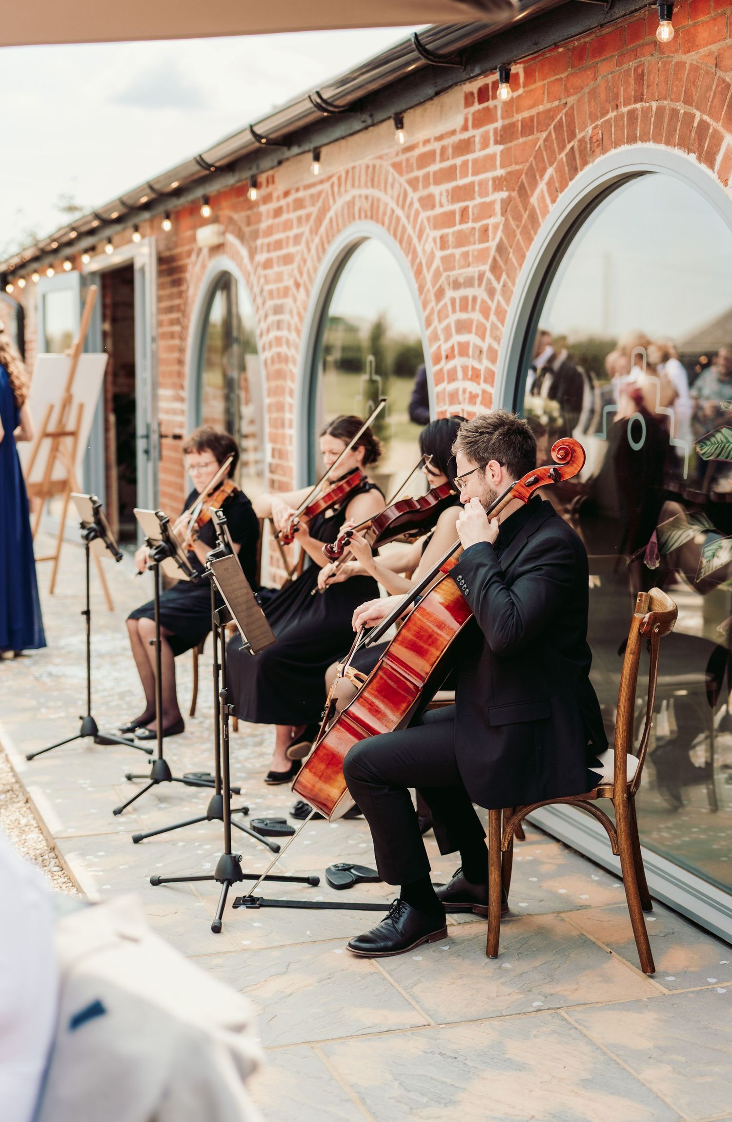 String quartet musicians in black attire perform in front of a brick wall at an outdoor venue.