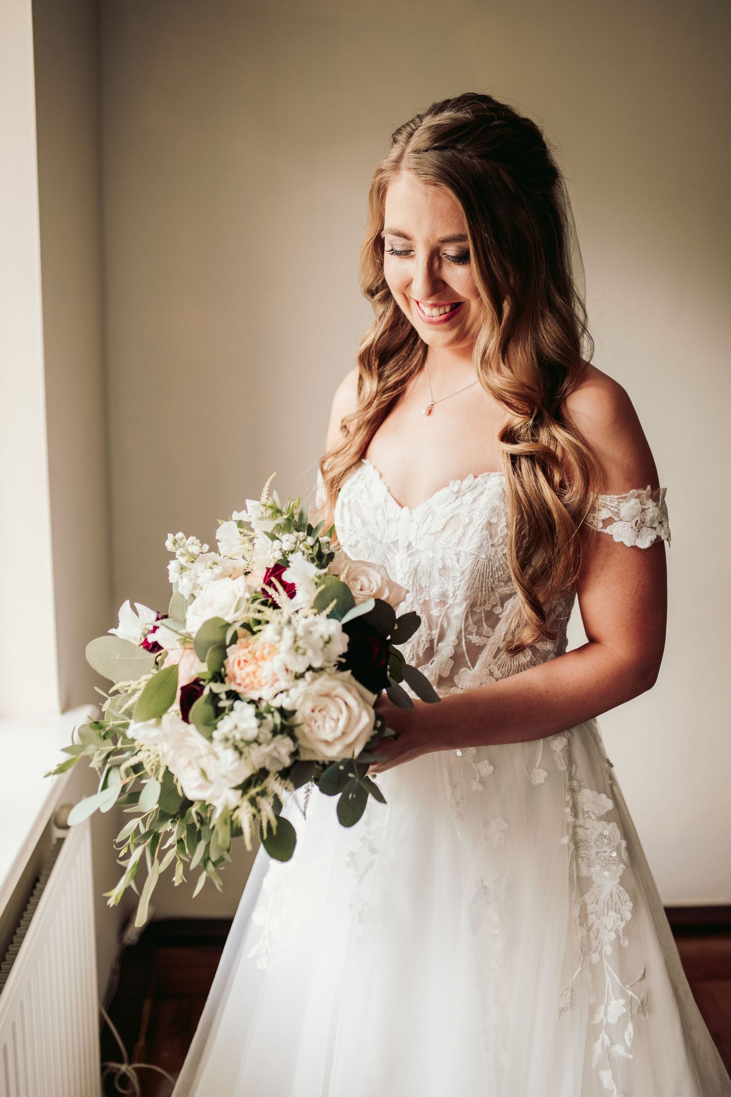 Beautiful bride in an off-shoulder lace wedding gown smiles while holding elegant white and blush floral bouquet.