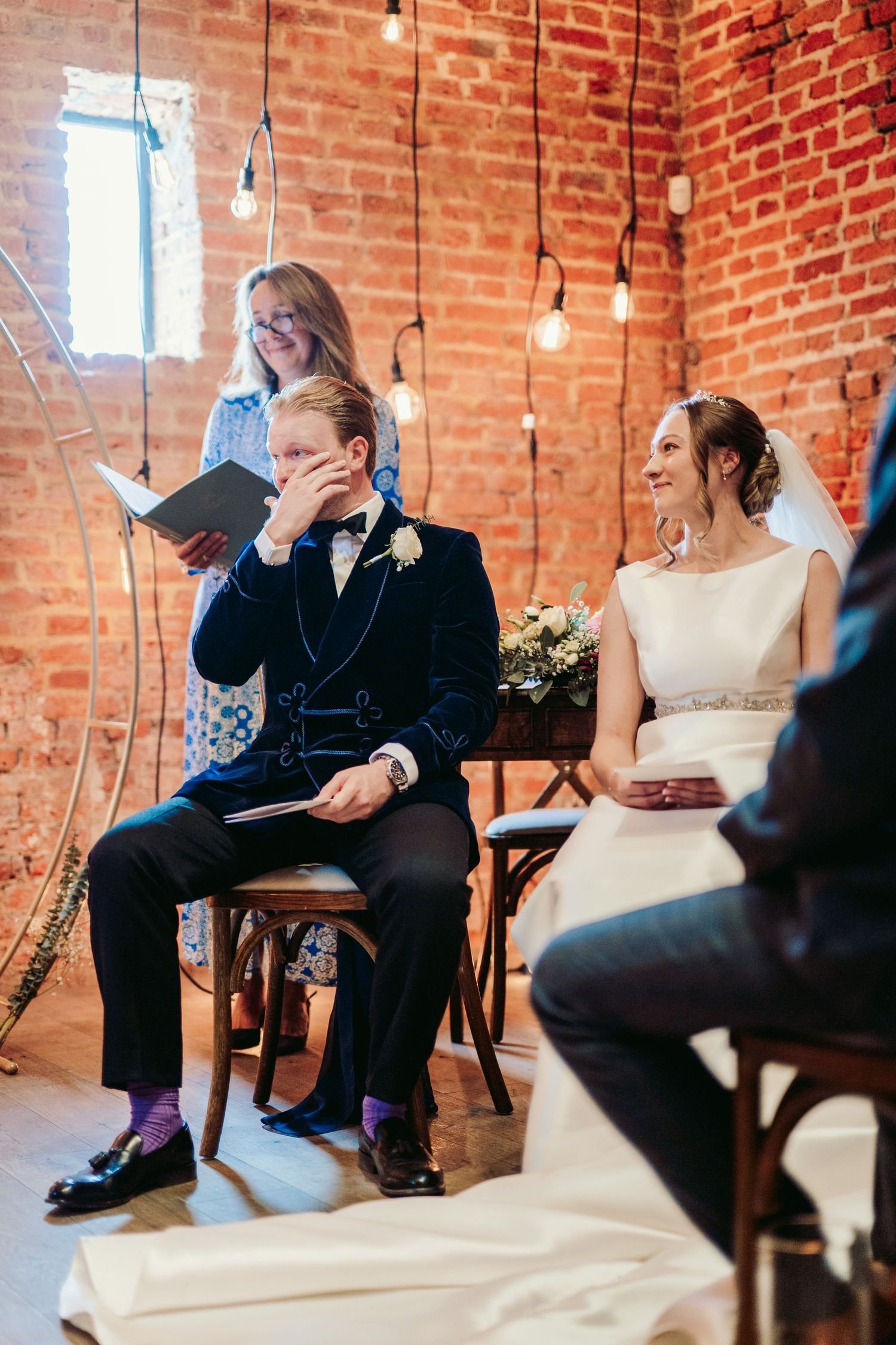 Emotional wedding ceremony in rustic brick venue with hanging Edison bulbs as couple shares laughter during the service.
