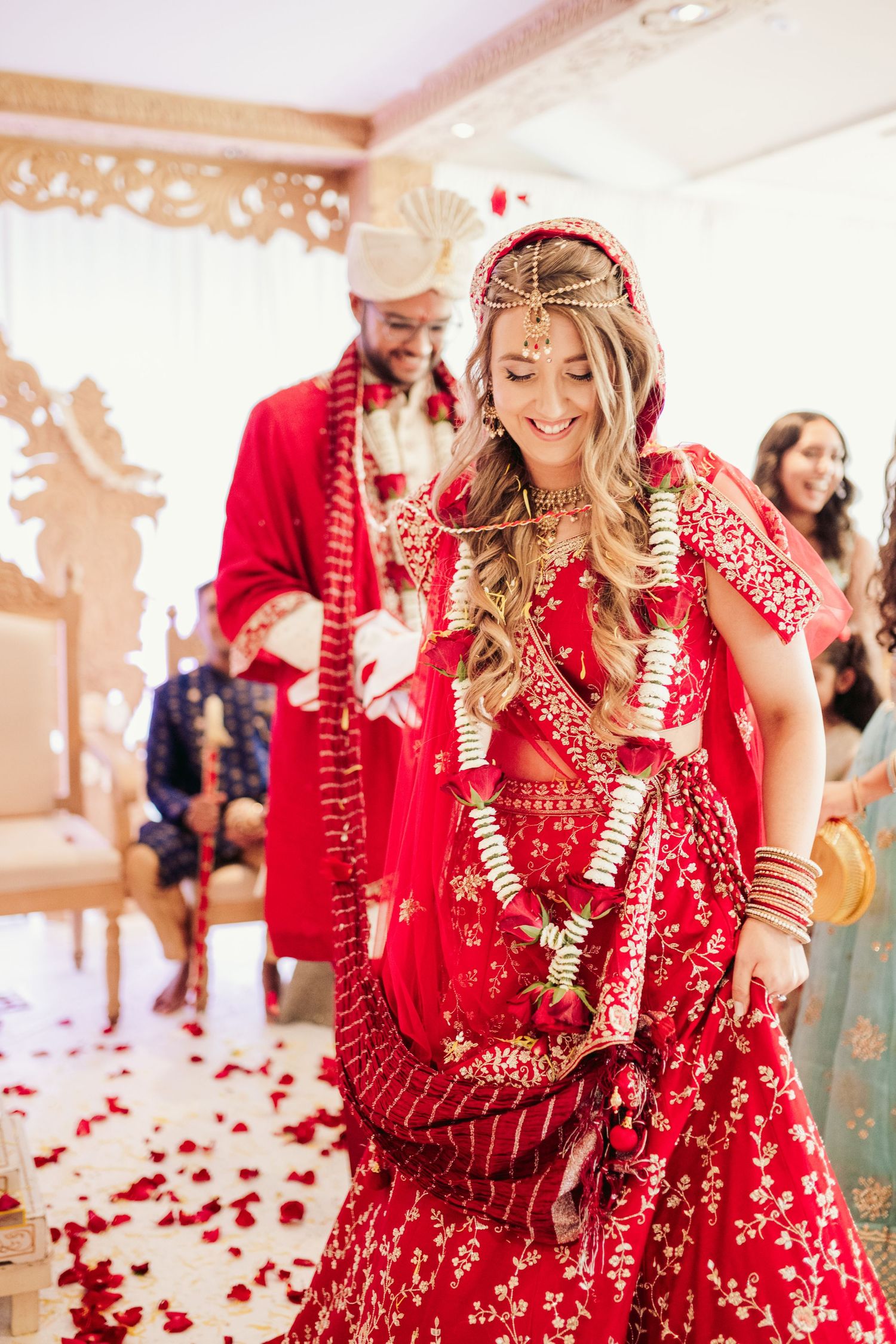 Two people in traditional red and gold Indian wedding attire dance and celebrate during a ceremony with rose petals.