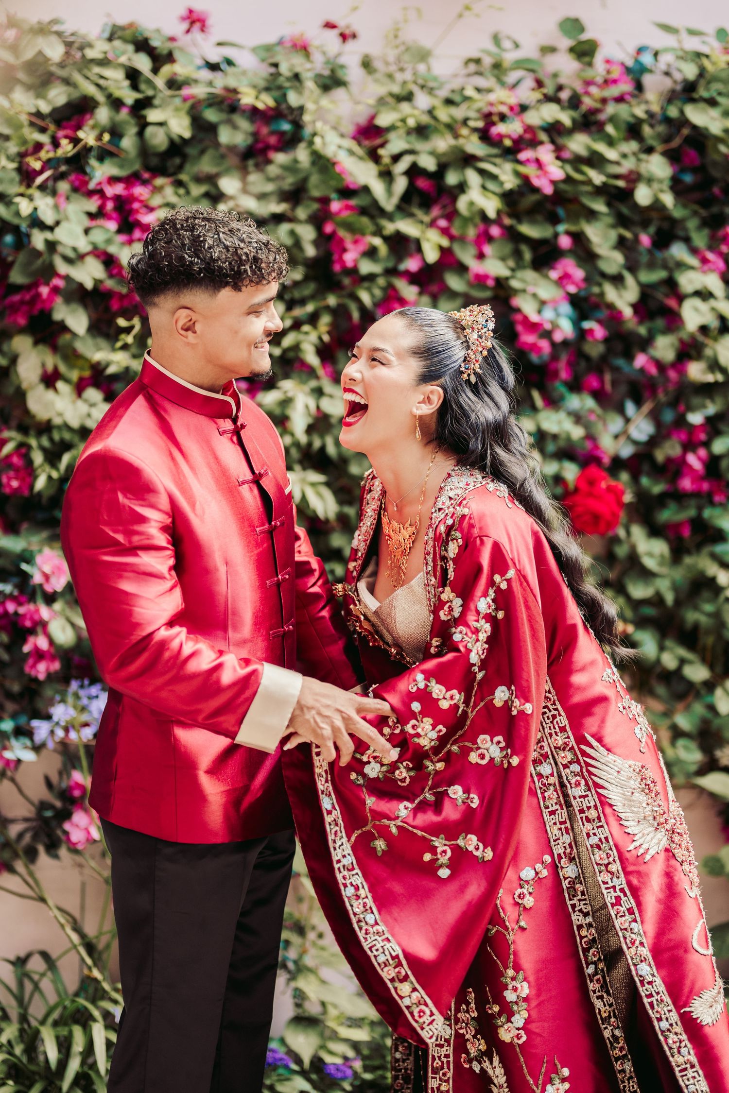 A couple in traditional red wedding attire shares a laugh in front of a flowering garden wall.