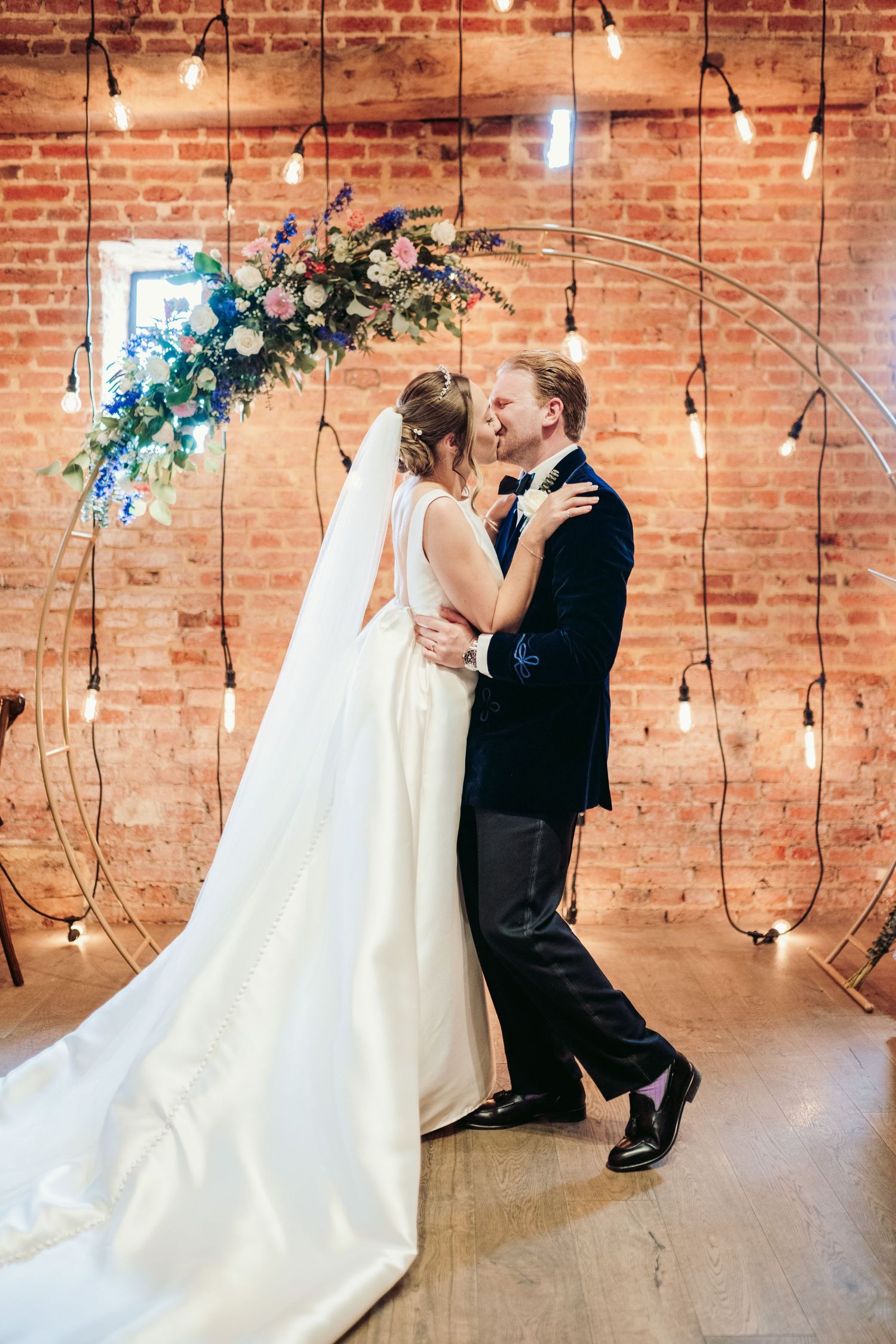 A romantic wedding moment captured in a rustic brick venue with hanging lights and floral decorations.