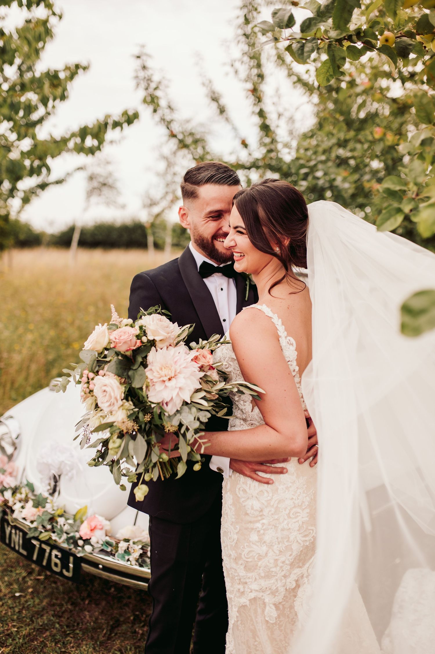 Romantic wedding portrait with couple embracing near vintage car with luxurious floral bouquet.