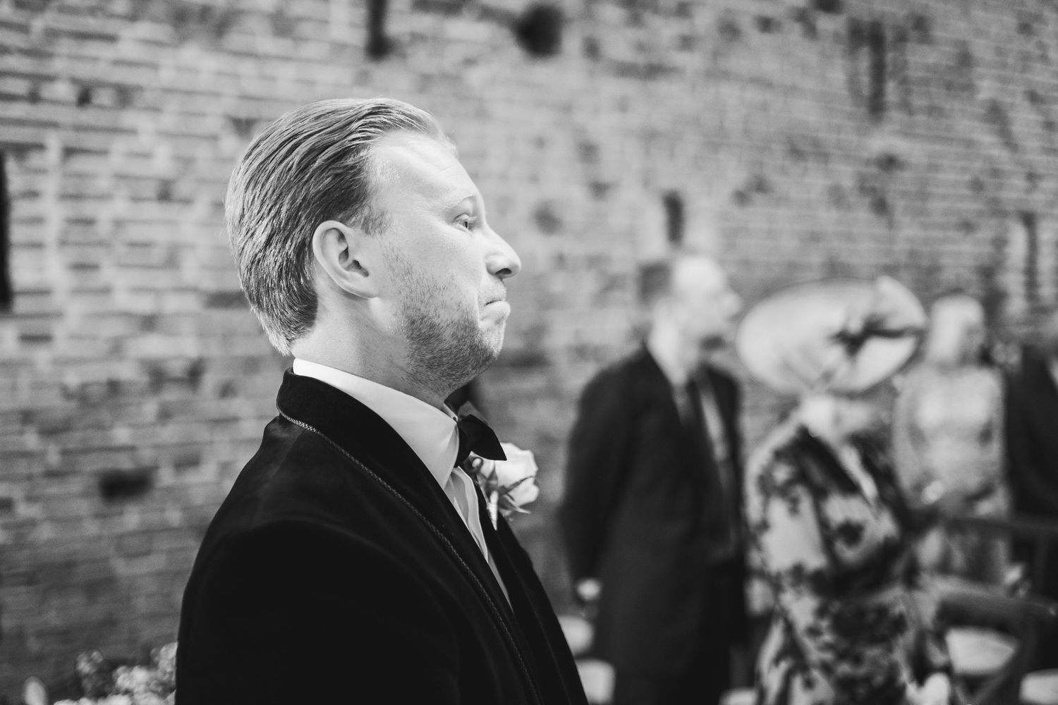 Black and white photo of a person in a formal suit with boutonniere standing in profile against a brick wall background.