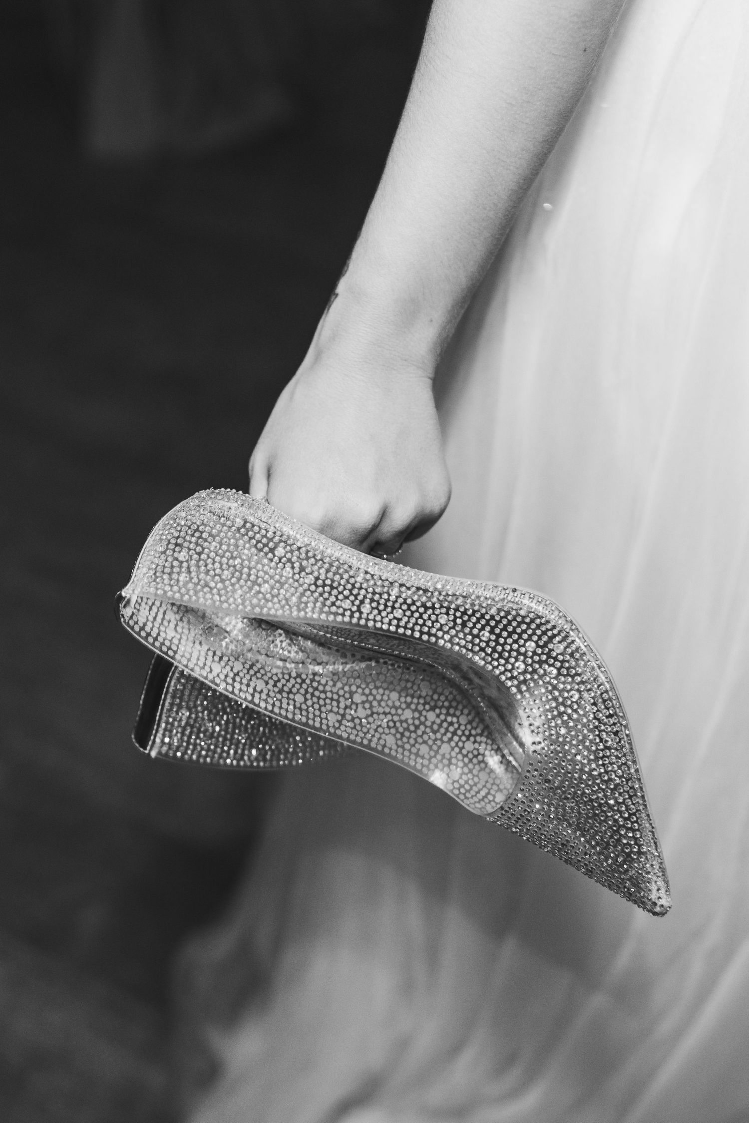 A sparkly crystal-encrusted high heel shoe being held against a white dress background in black and white.