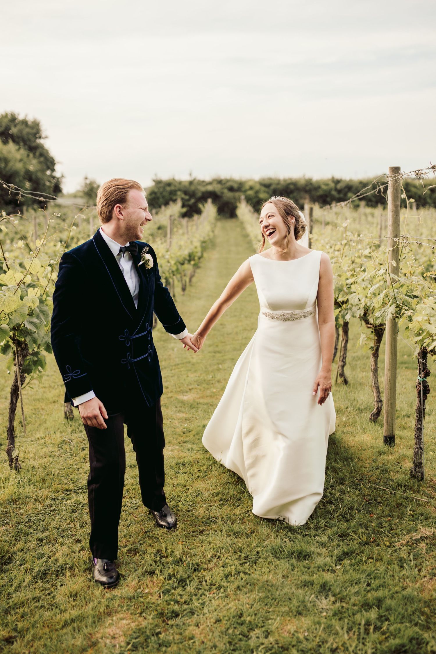 A couple shares a joyful moment walking through a vineyard in wedding attire.