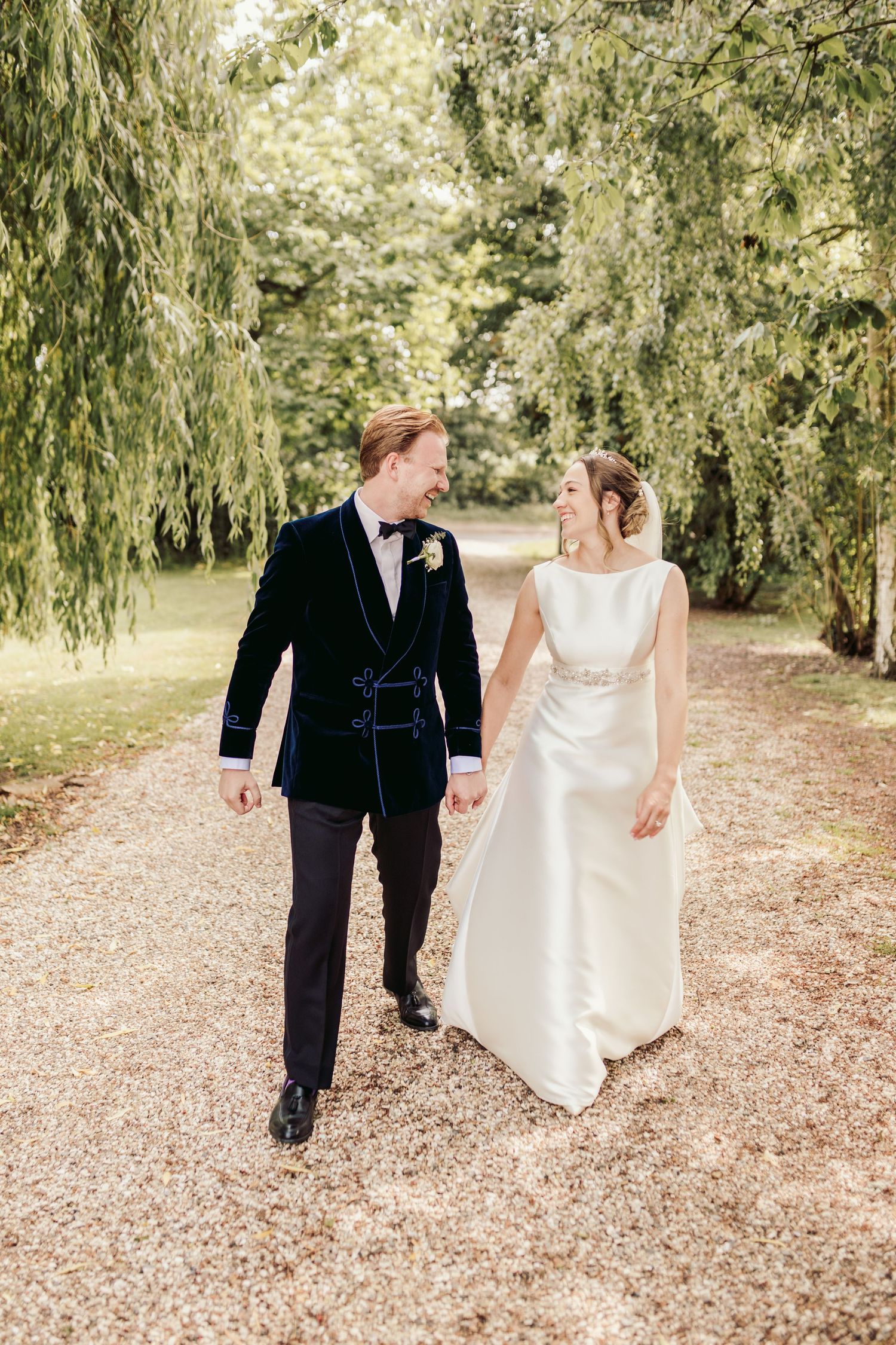 Newlywed couple walking together on a leaf-covered path under willow trees in a romantic garden setting.