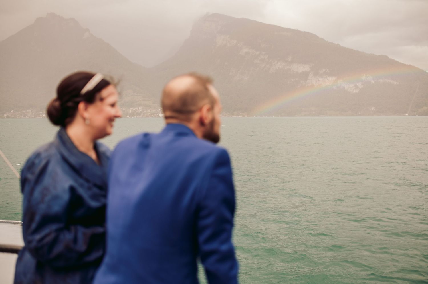 Two people stand on a boat deck overlooking misty mountains and turquoise waters on a cloudy day. Spiez switzerland destination weddingSWITZ