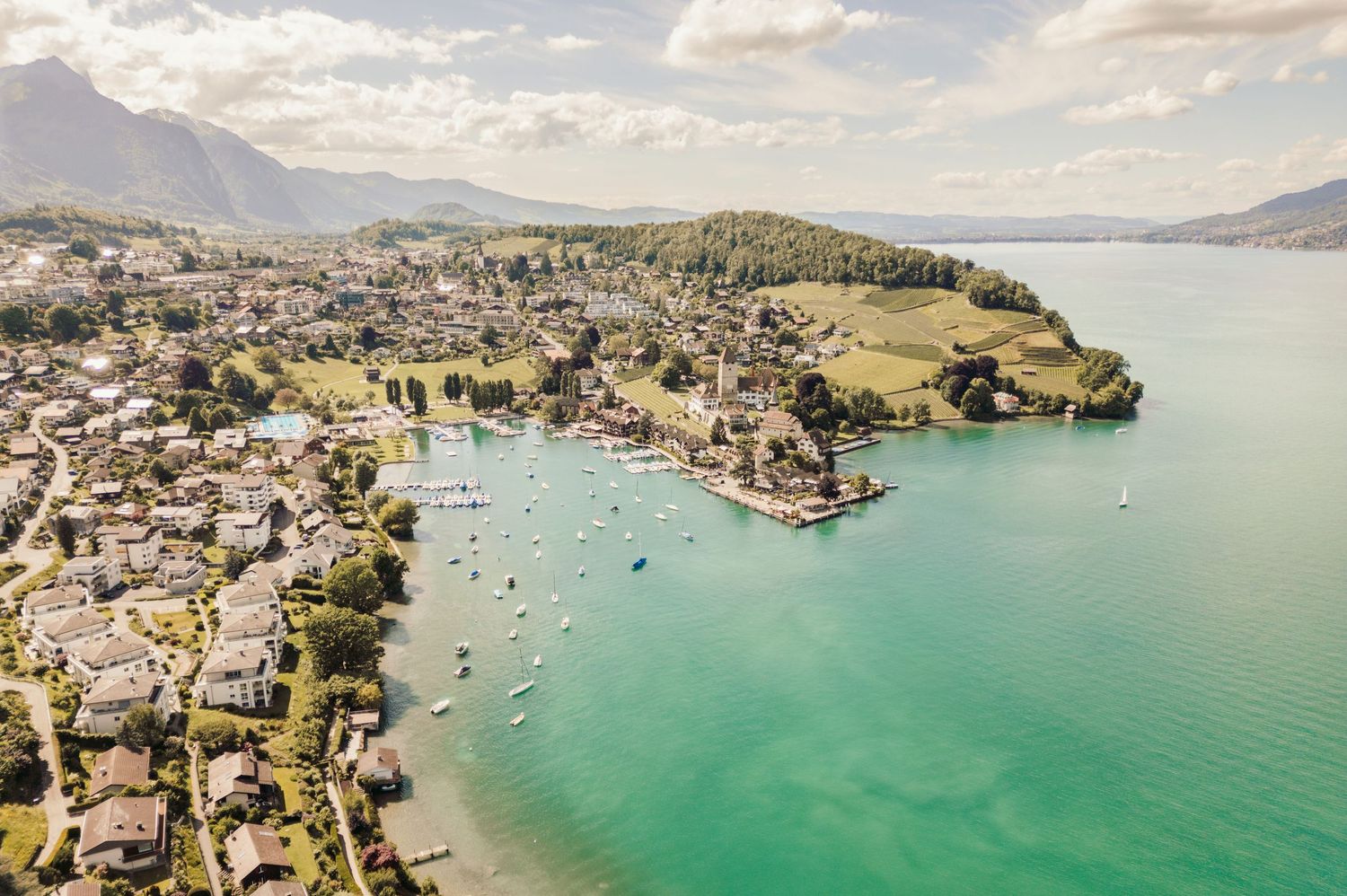 Aerial view of a coastal town in Switzerland with turquoise waters, boats in harbor, and mountains in background.Spiez switzerland