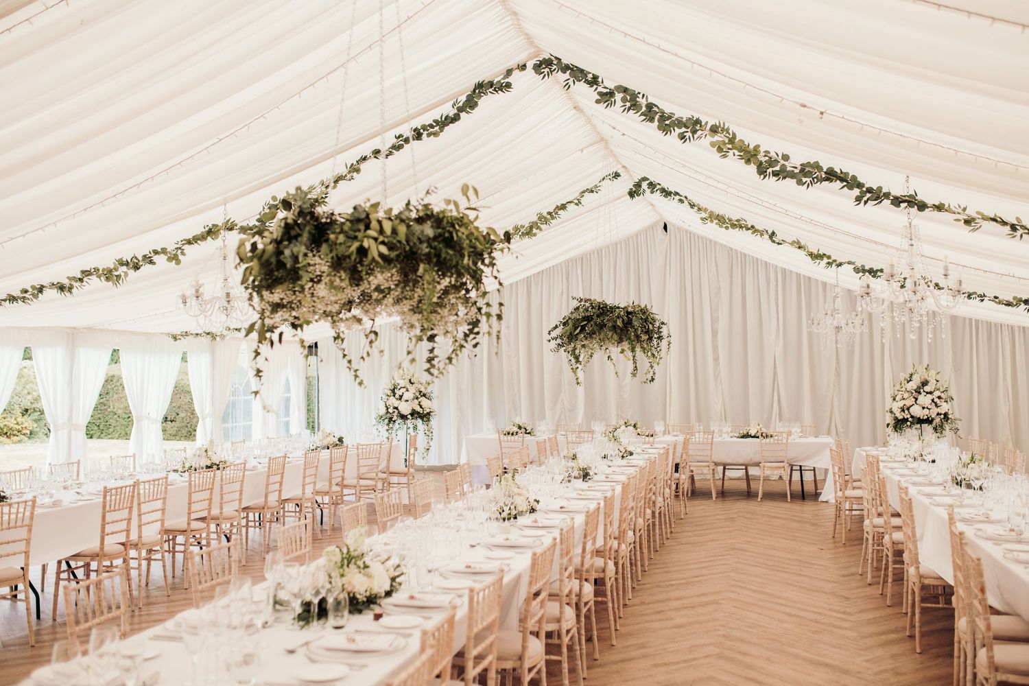 Elegant white wedding tent interior with draped fabric ceiling, floral chandeliers and long banquet tables set for reception.