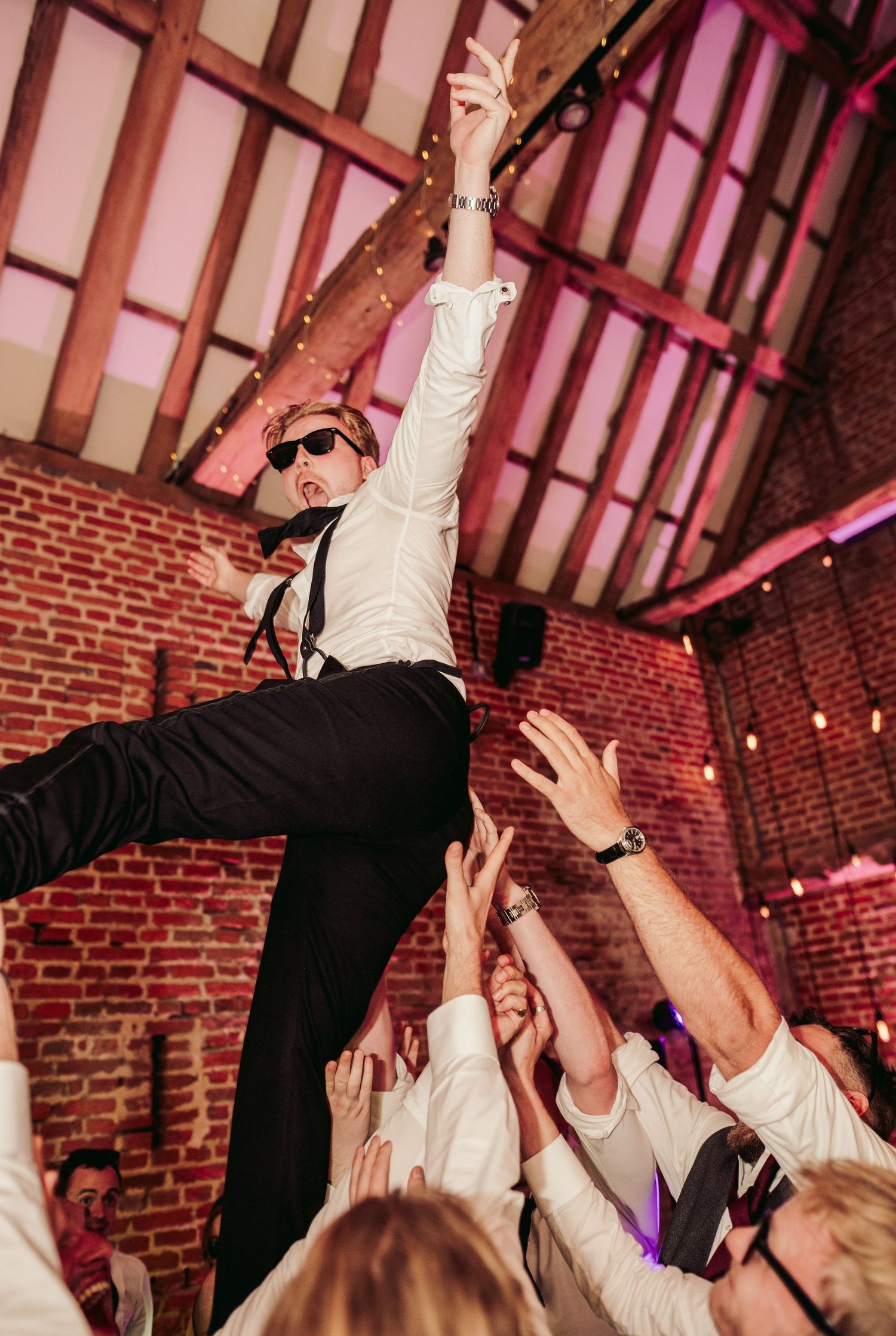 Wedding guests lift and carry someone across a dance floor at a reception in a rustic brick venue with pink lighting.