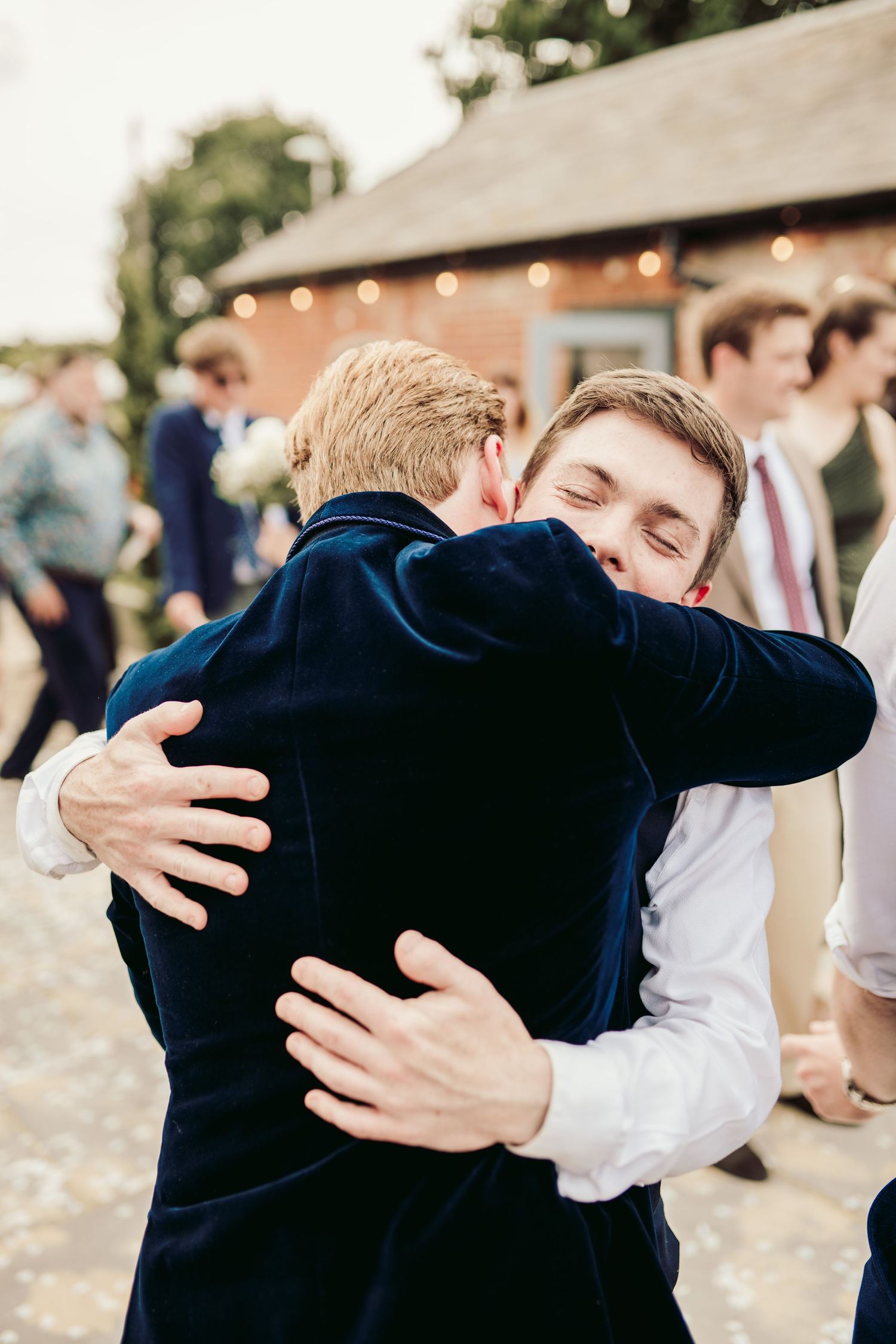 Two people share an emotional embrace at an outdoor event with string lights and guests visible in the background.