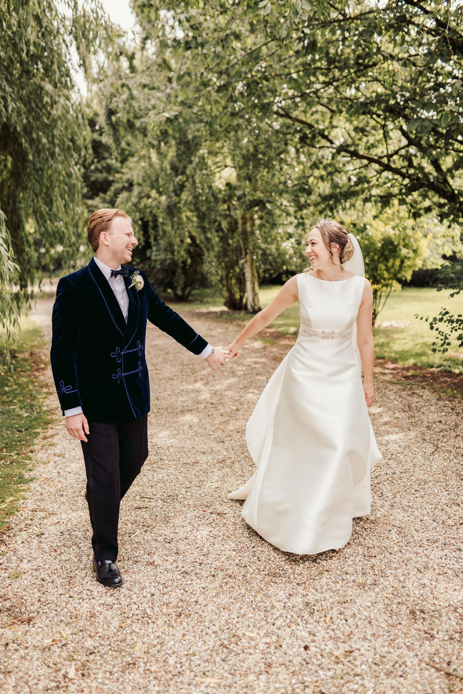 A couple in wedding attire playfully interacts while walking on a gravel path surrounded by lush greenery.