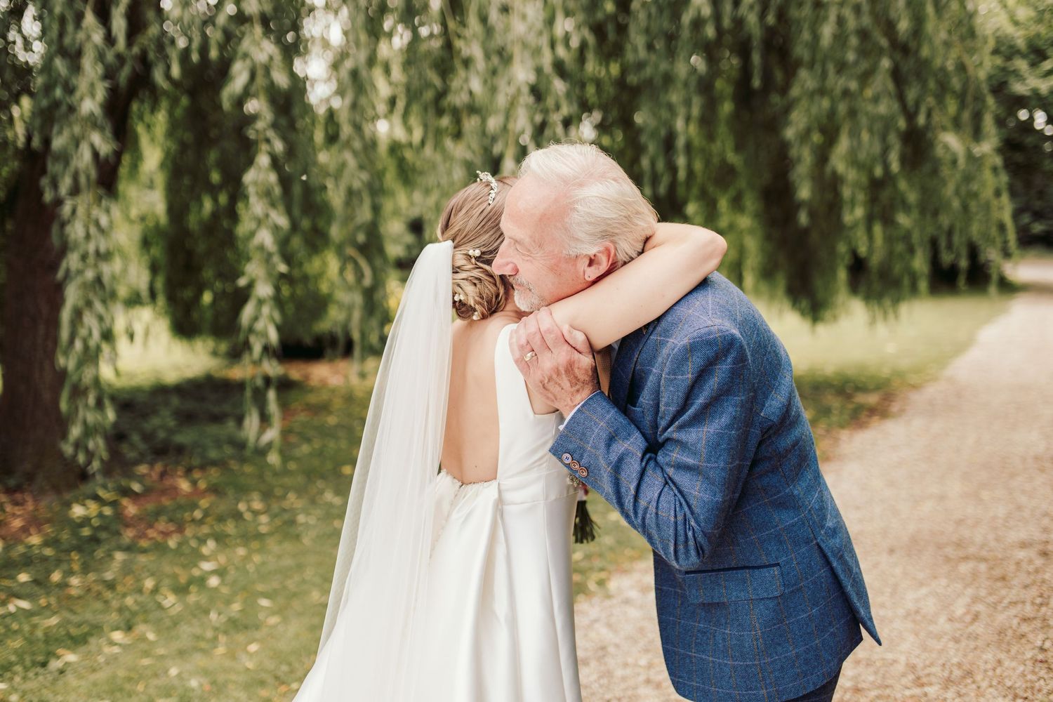 An emotional moment between a bride in a white veil and an elderly person in a blue suit under a weeping willow tree.