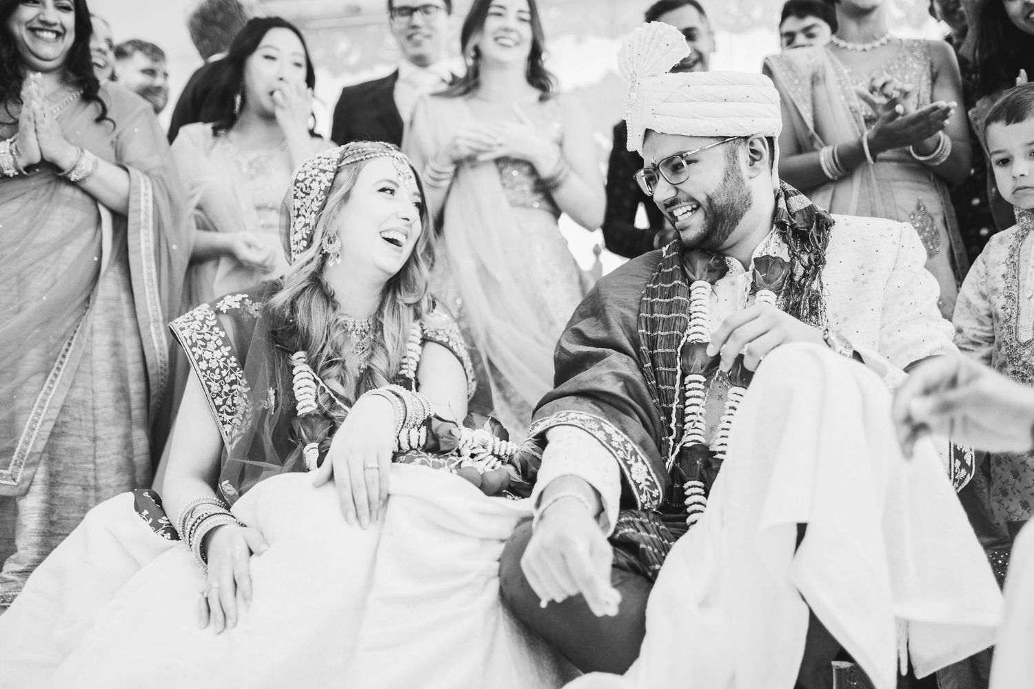 A black and white image shows seated wedding guests smiling and clapping at a celebration together.