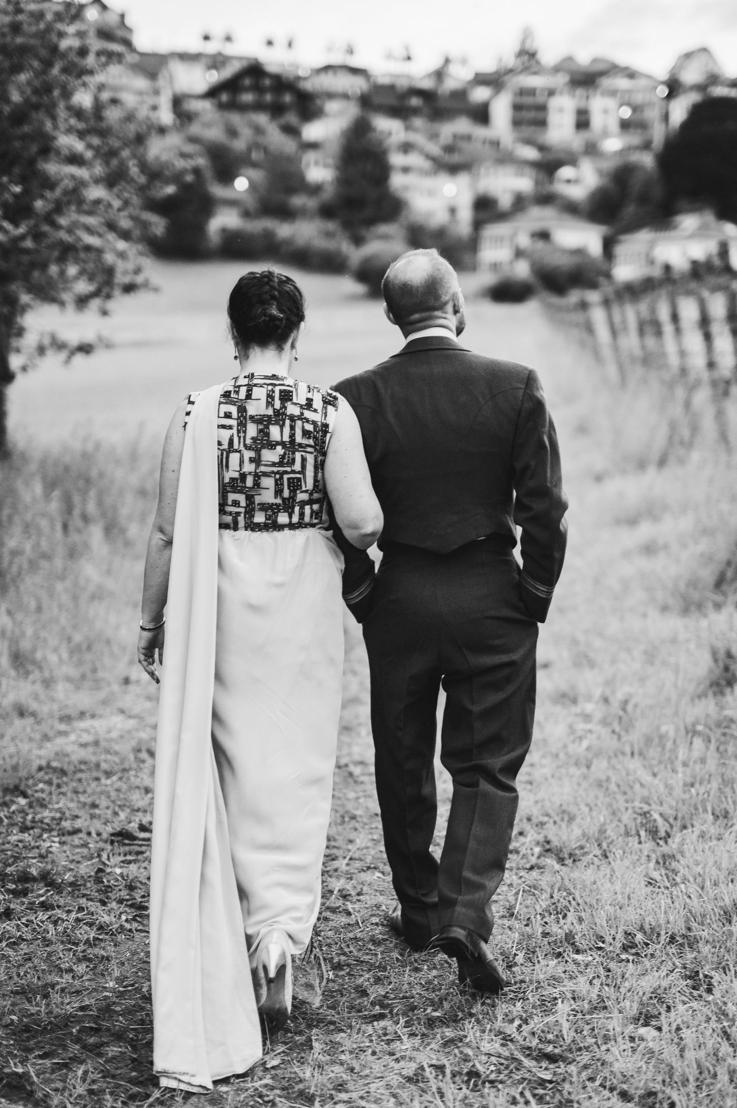 A black and white photo of a couple walking up a grassy hill toward houses in the distance.Spiez switzerland destination wedding