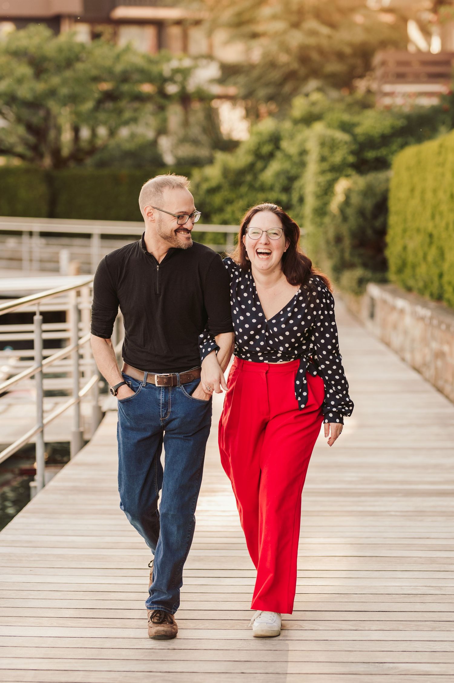 A couple walking together and laughing on a wooden boardwalk, wearing a black top and polka dots with bright red pants.Spiez switzerland