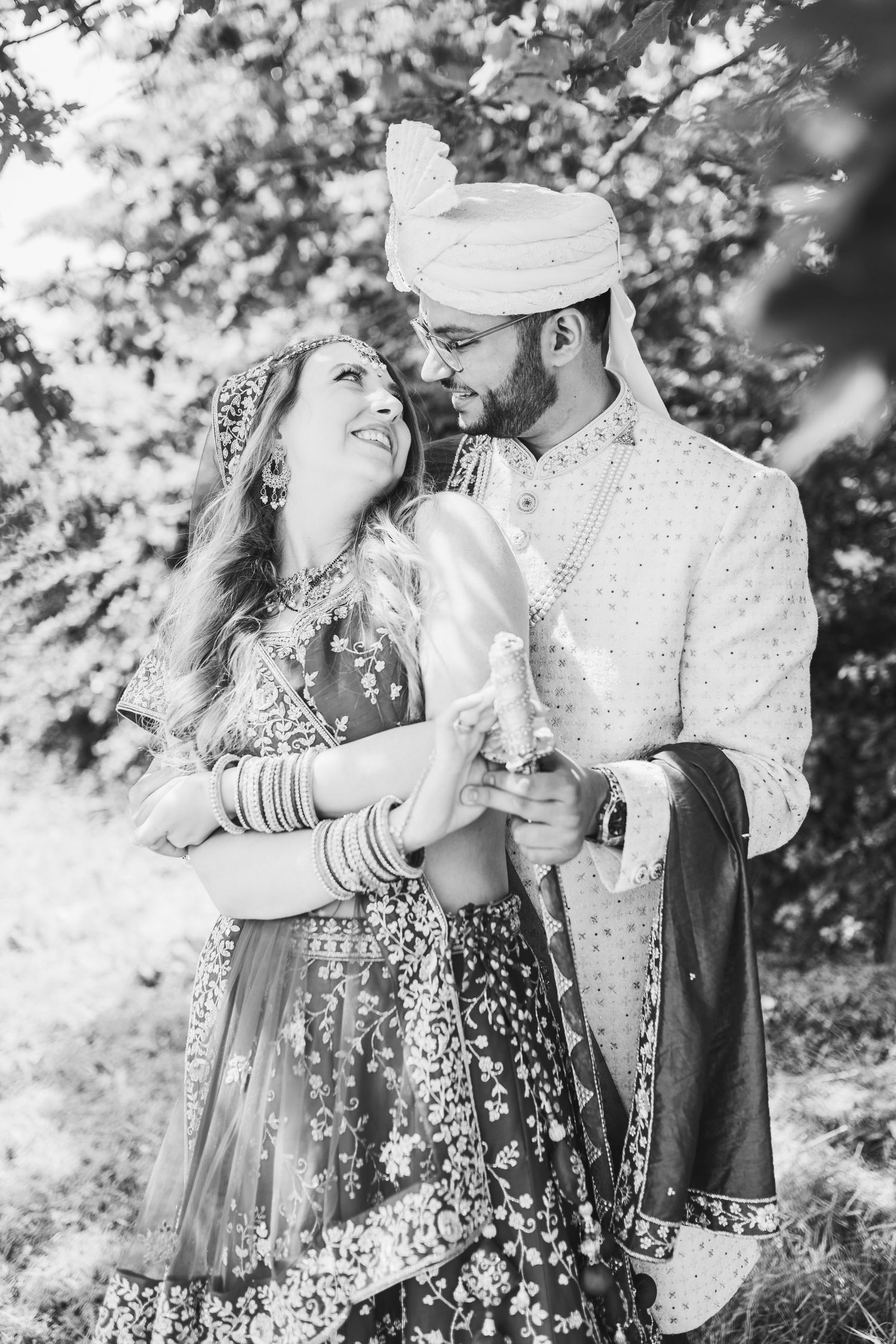 A couple in traditional South Asian wedding attire embrace outdoors in a romantic black and white photograph.