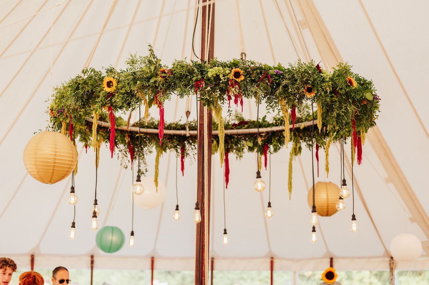 Floral chandelier with hanging lanterns and greenery decorates a wedding tent ceiling.