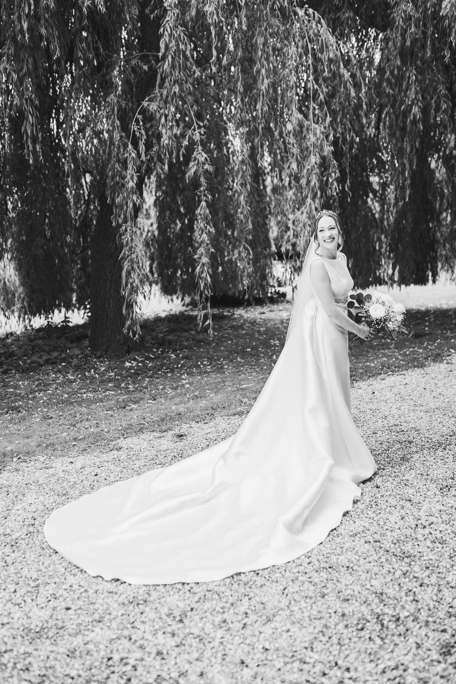 A bride in a flowing white wedding gown with a long cathedral train stands under weeping willow trees.