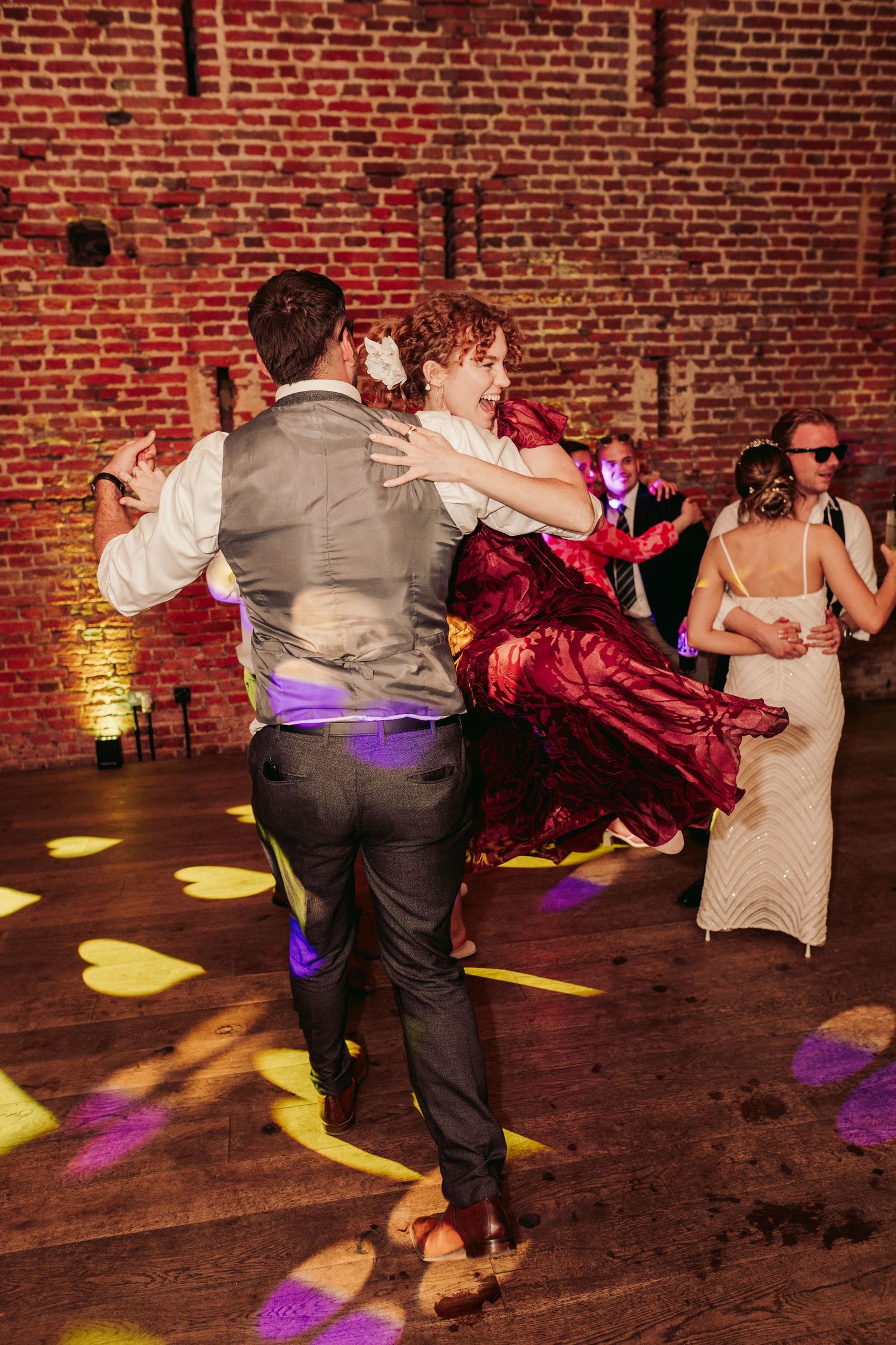 Dancers twirl and spin on a wooden dance floor against exposed brick walls illuminated by colorful spotlights.