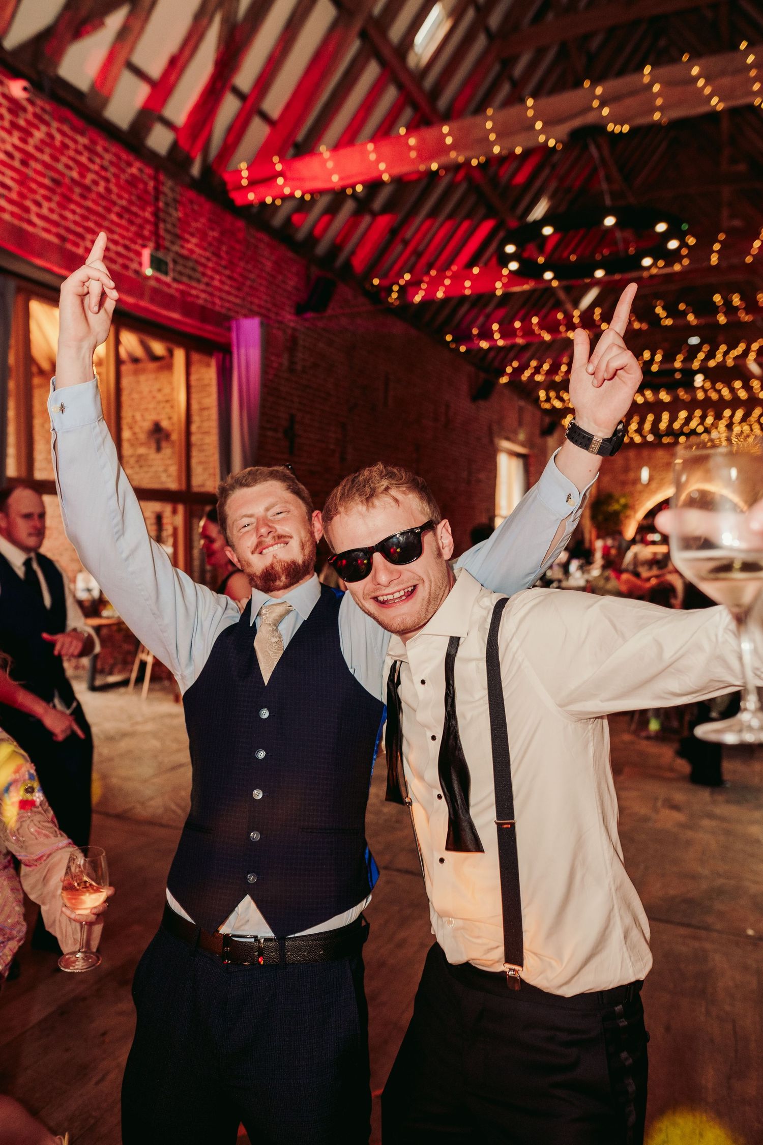 Wedding guests dancing and celebrating with raised arms in a rustic barn venue with red ambient lighting and string lights.
