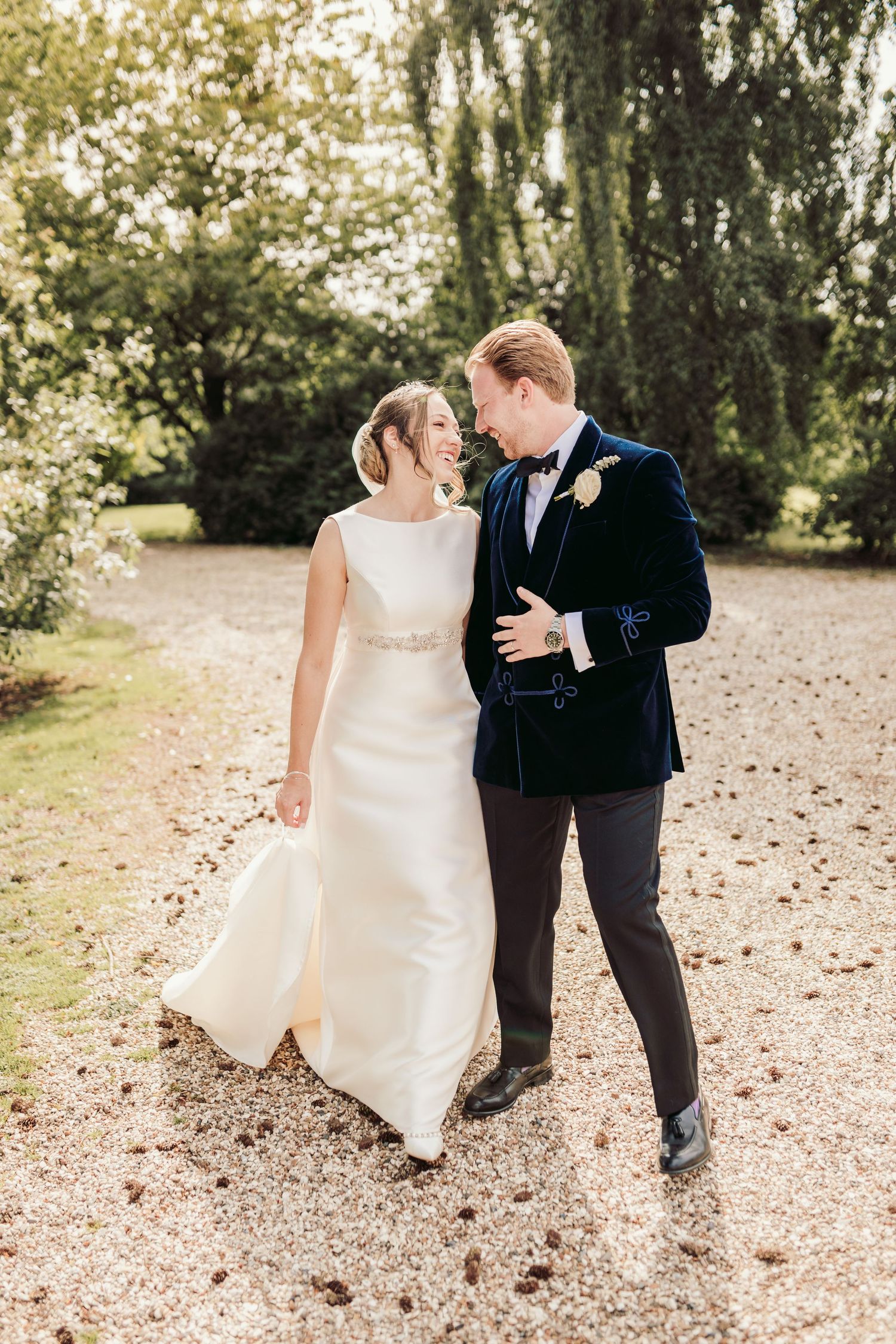 A romantic couple in wedding attire share a playful moment on a leaf-covered path in a wooded setting.