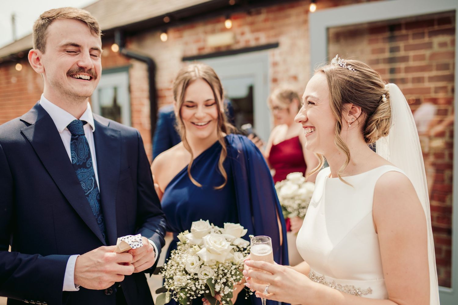A group of wedding guests and wedding party members celebrate during an outdoor reception at a brick venue.