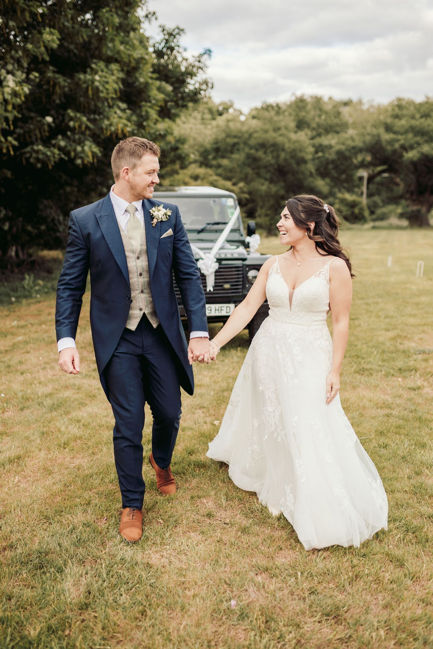 Newlyweds walk playfully through grassy field in formal wedding attire.