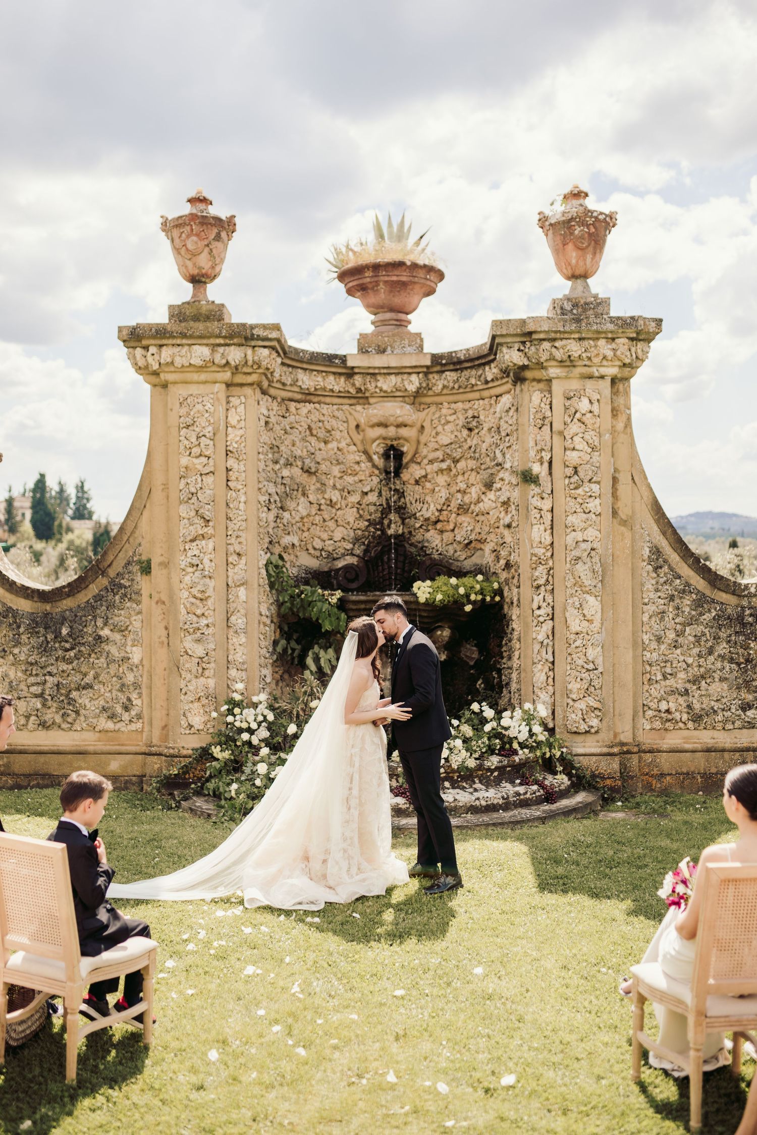 Wedding ceremony in front of ornate stone archway with decorative urns and flower arrangements.