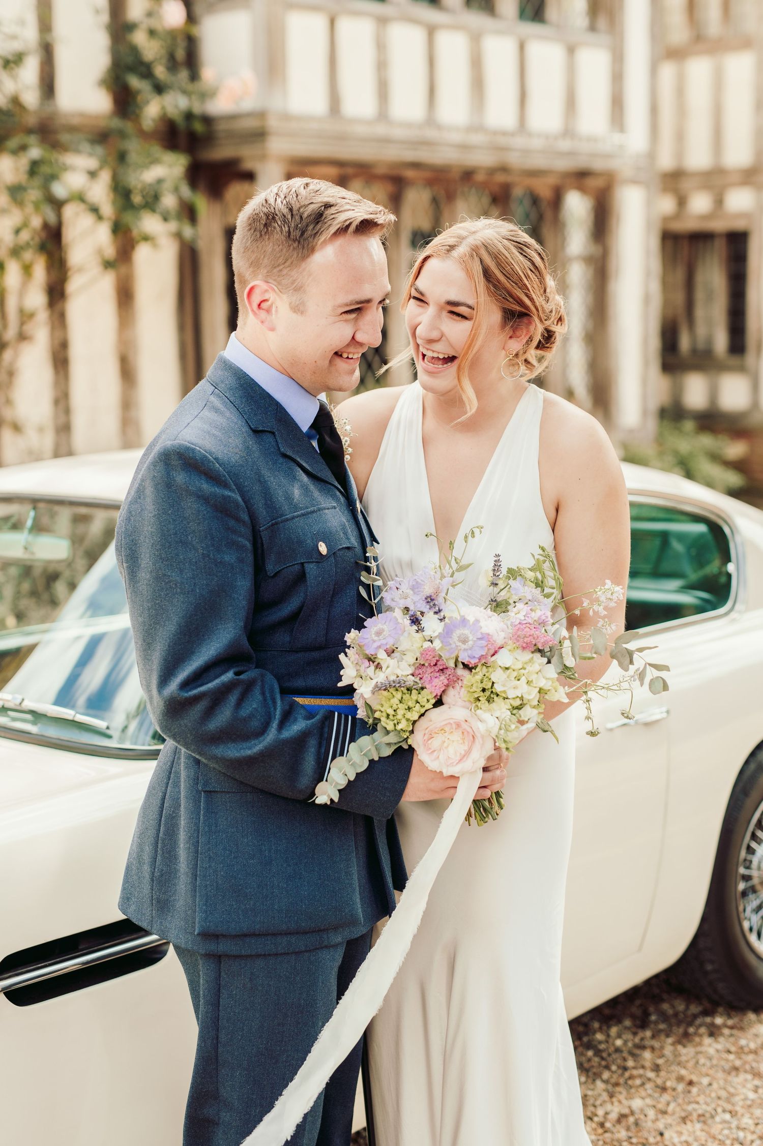 Newlyweds share intimate moment while leaning against vintage white car.