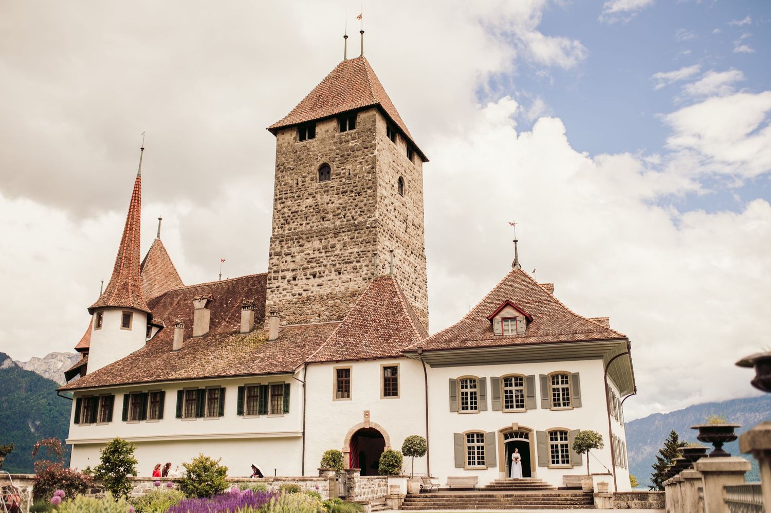 Historic stone castle with tall tower and white facades stands atop hill against cloudy mountain backdrop in Switzerland. Spiez switzerland