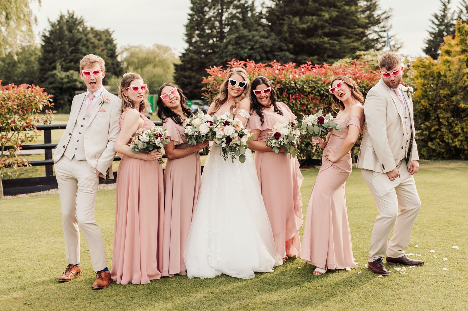 Wedding party poses together outdoors in blush pink and beige attire on a sunny garden lawn.